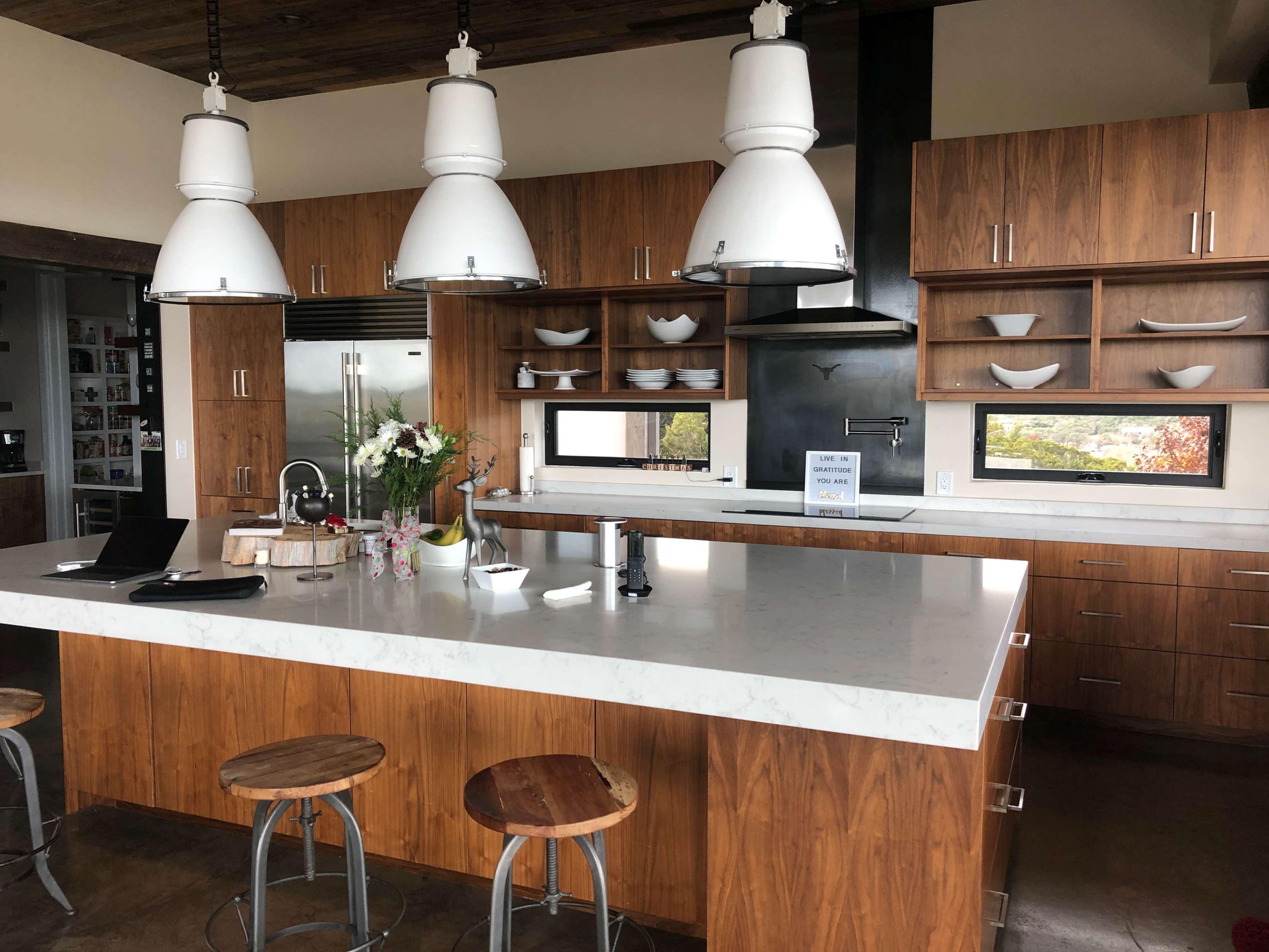 A modern kitchen features a central island with bar stools, wooden cabinetry, stainless steel appliances, and pendant lights hanging above.