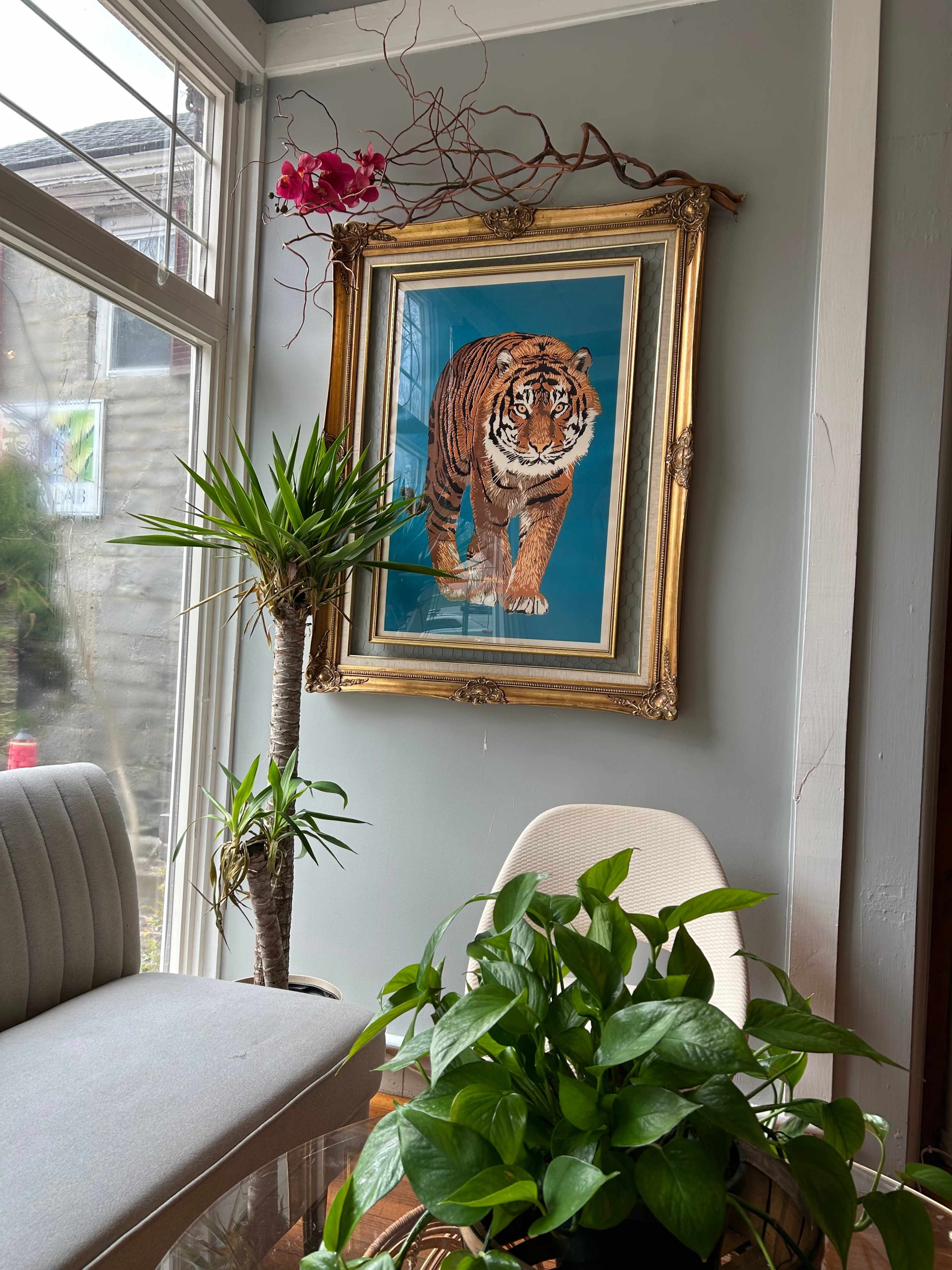 A large framed painting of a tiger hangs on a gray wall alongside a potted plant and a small decorative tree, all situated in a cozy seating area.