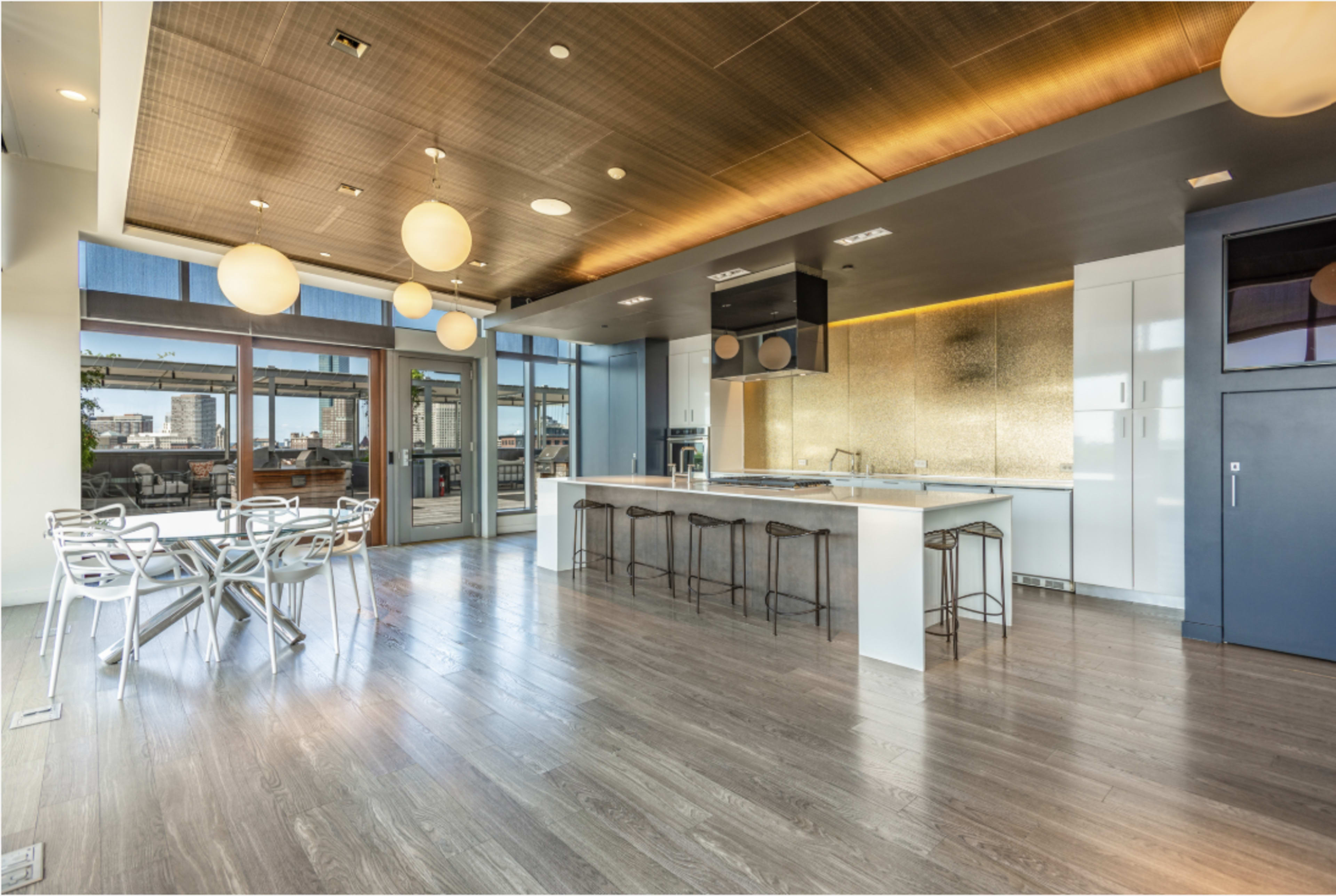 The image shows a modern kitchen with a large island, metallic backsplash, and a dining area featuring a glass table and four chairs.
