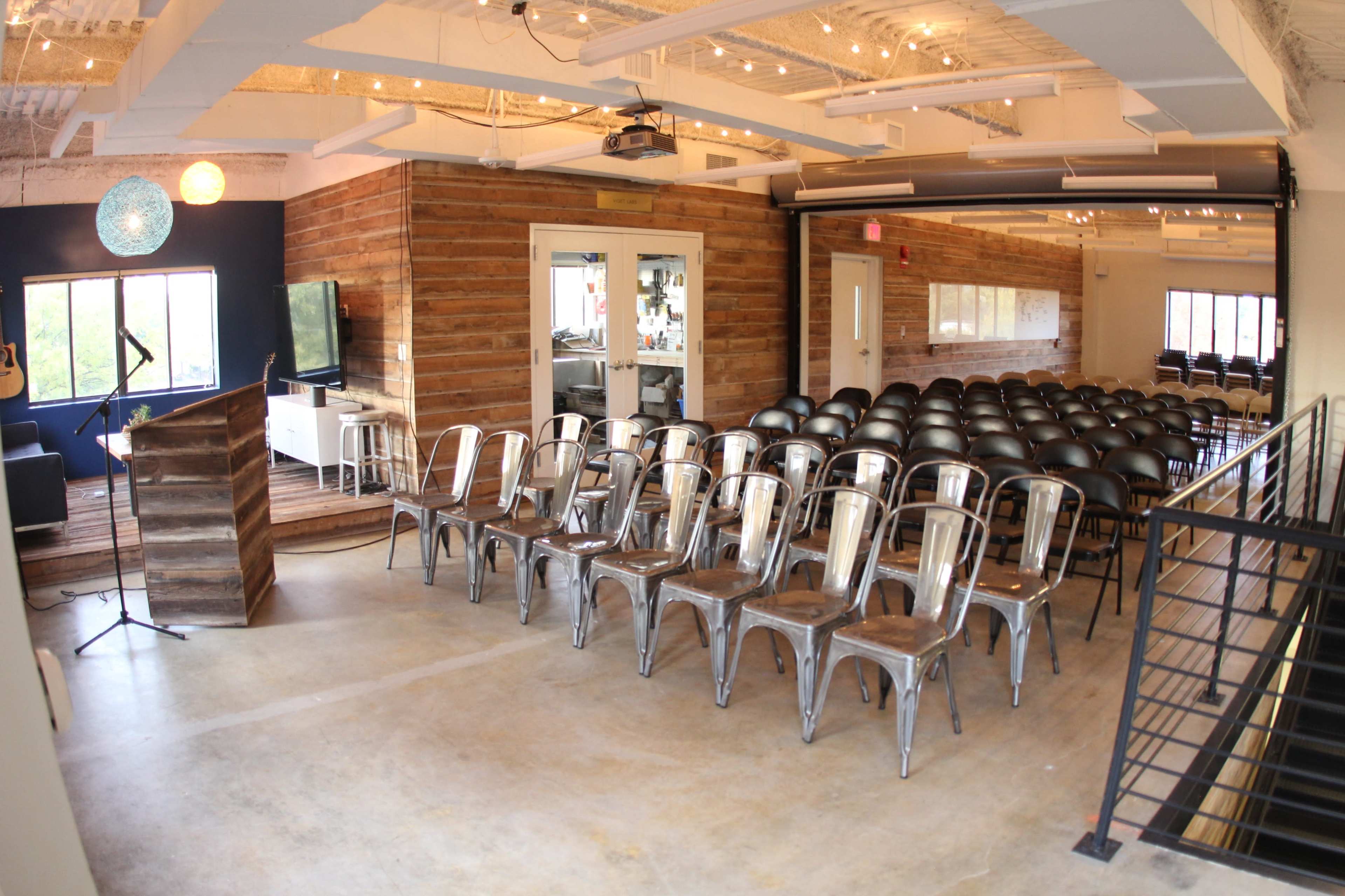 A room with a wooden stage and rows of metal chairs set up for an event, featuring large windows and modern decor.