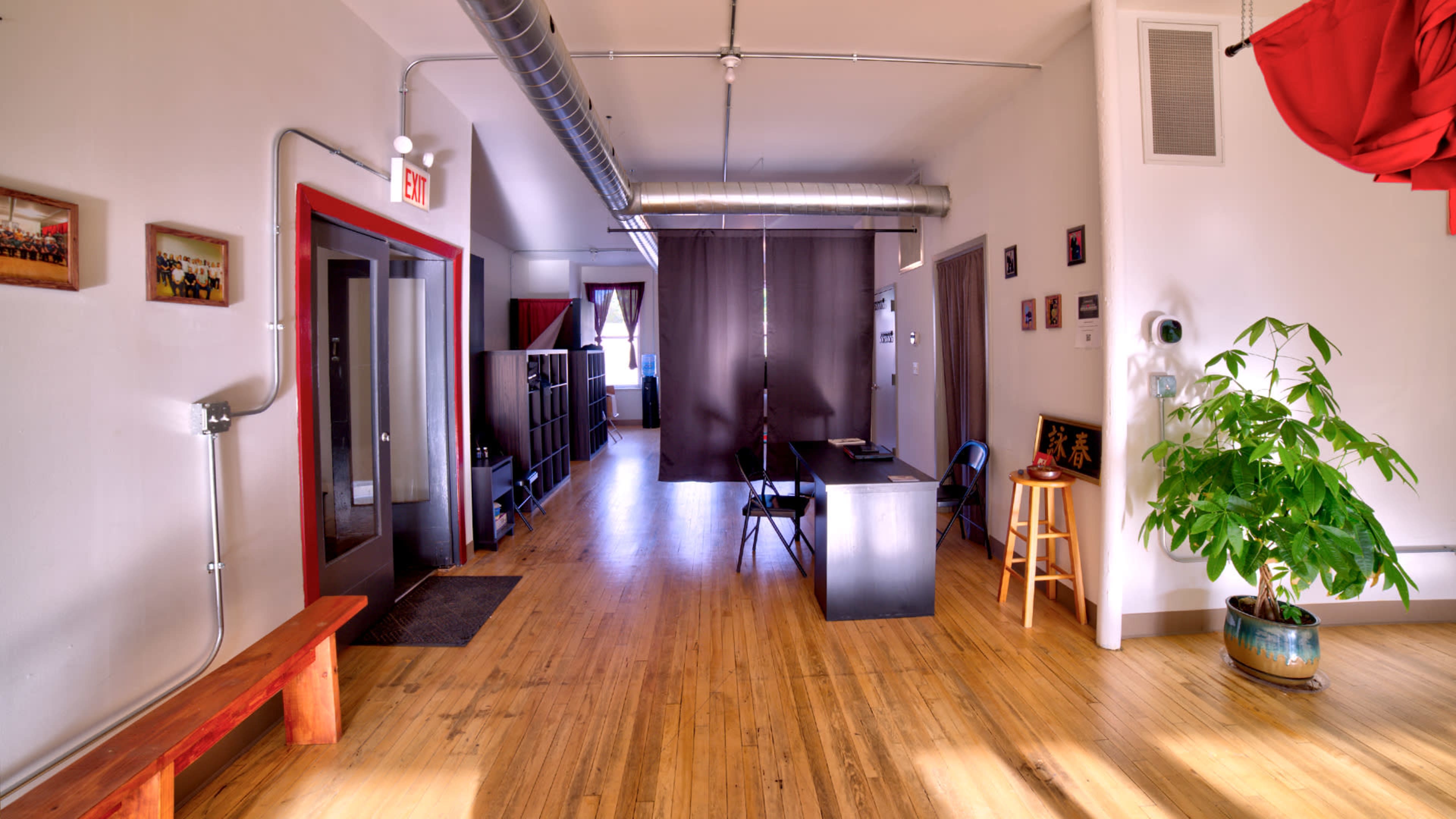 The image shows a spacious interior room with wooden flooring, featuring a reception area with a desk, chairs, and plants, along with a curtain partially dividing the space.