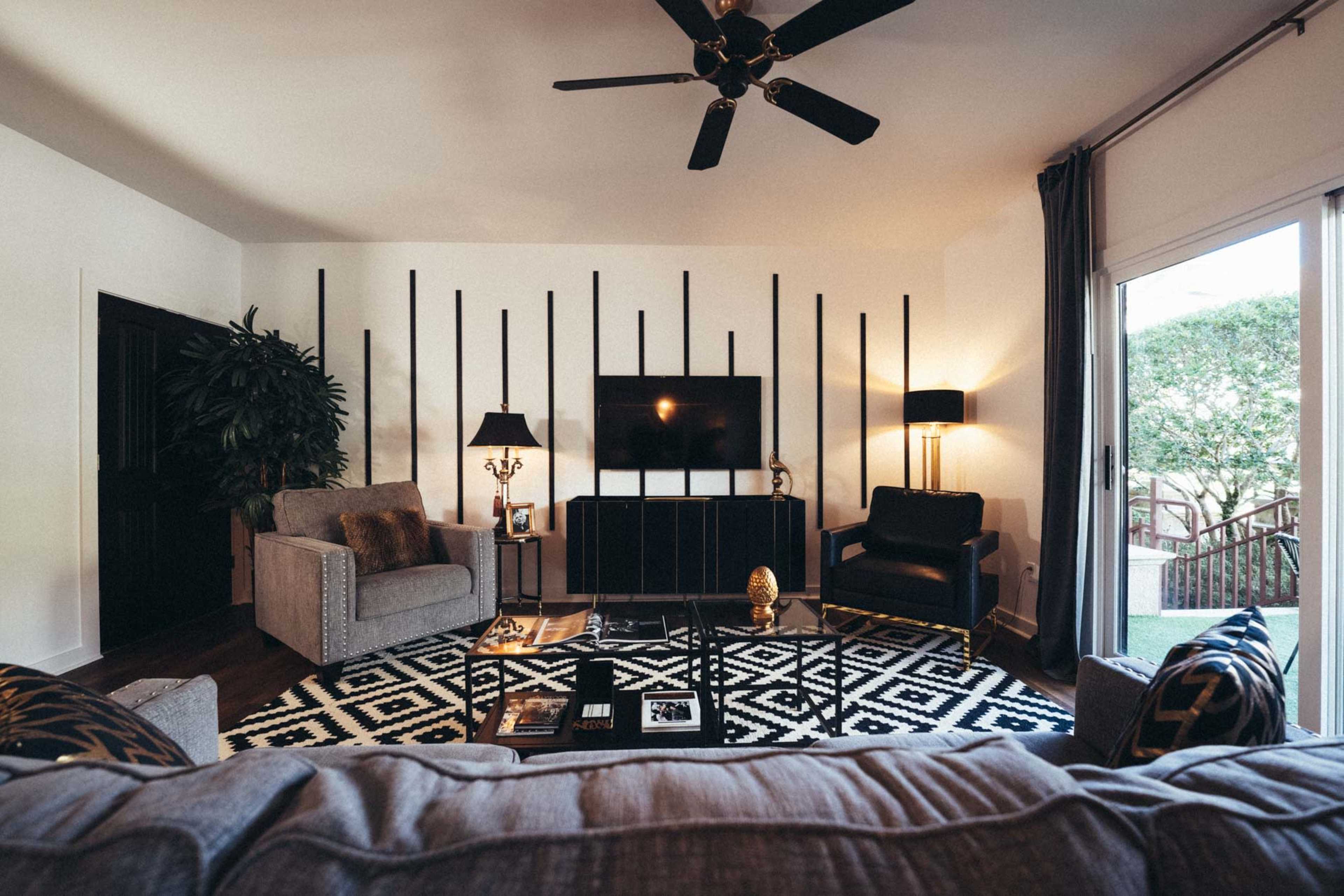 The living room features two sofas, a coffee table, a flat-screen TV mounted on the wall, and large windows with a view of the outdoors.
