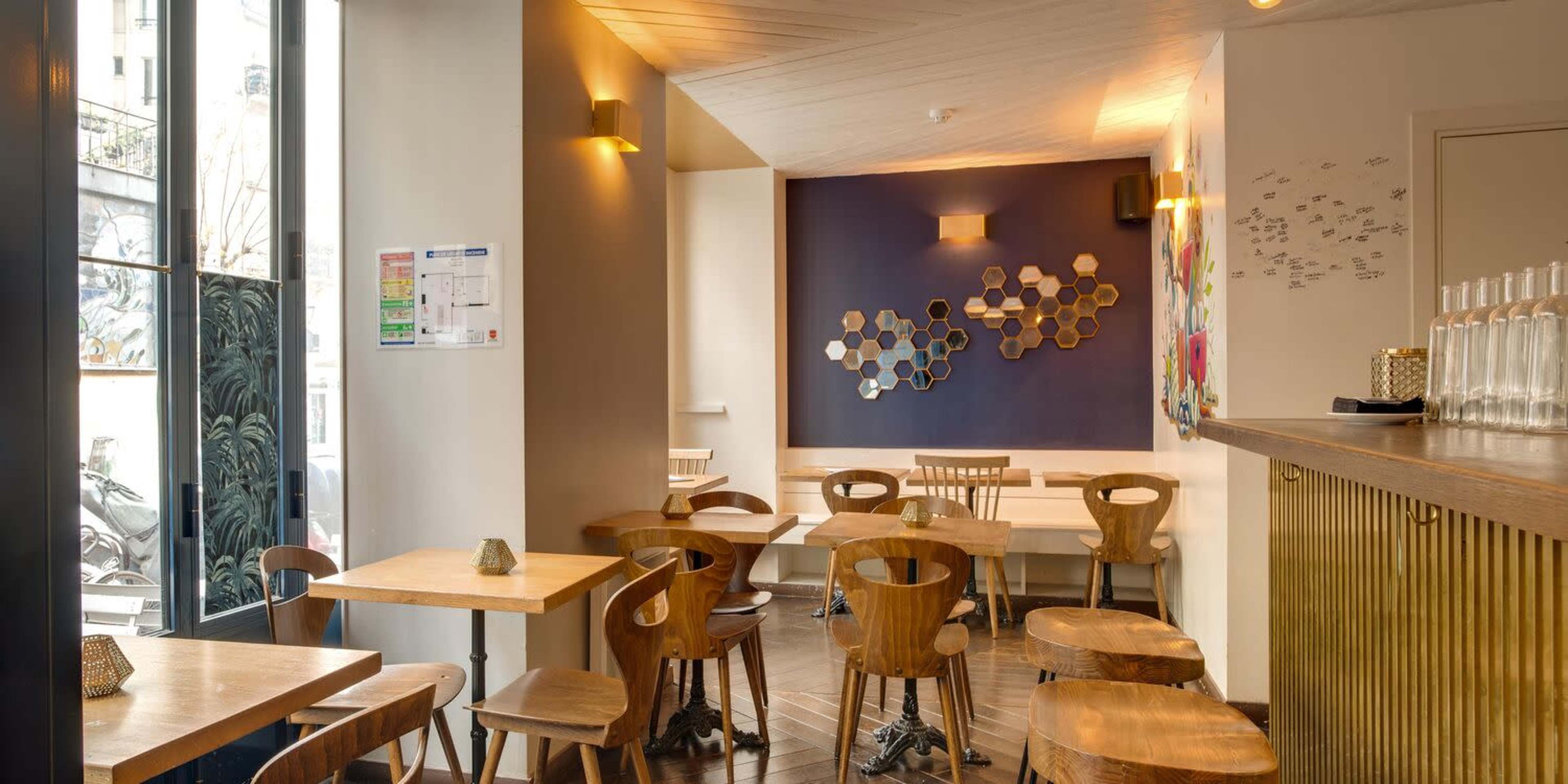A modern café interior with wooden tables and chairs, a colorful wall design, and a bar area on the right.