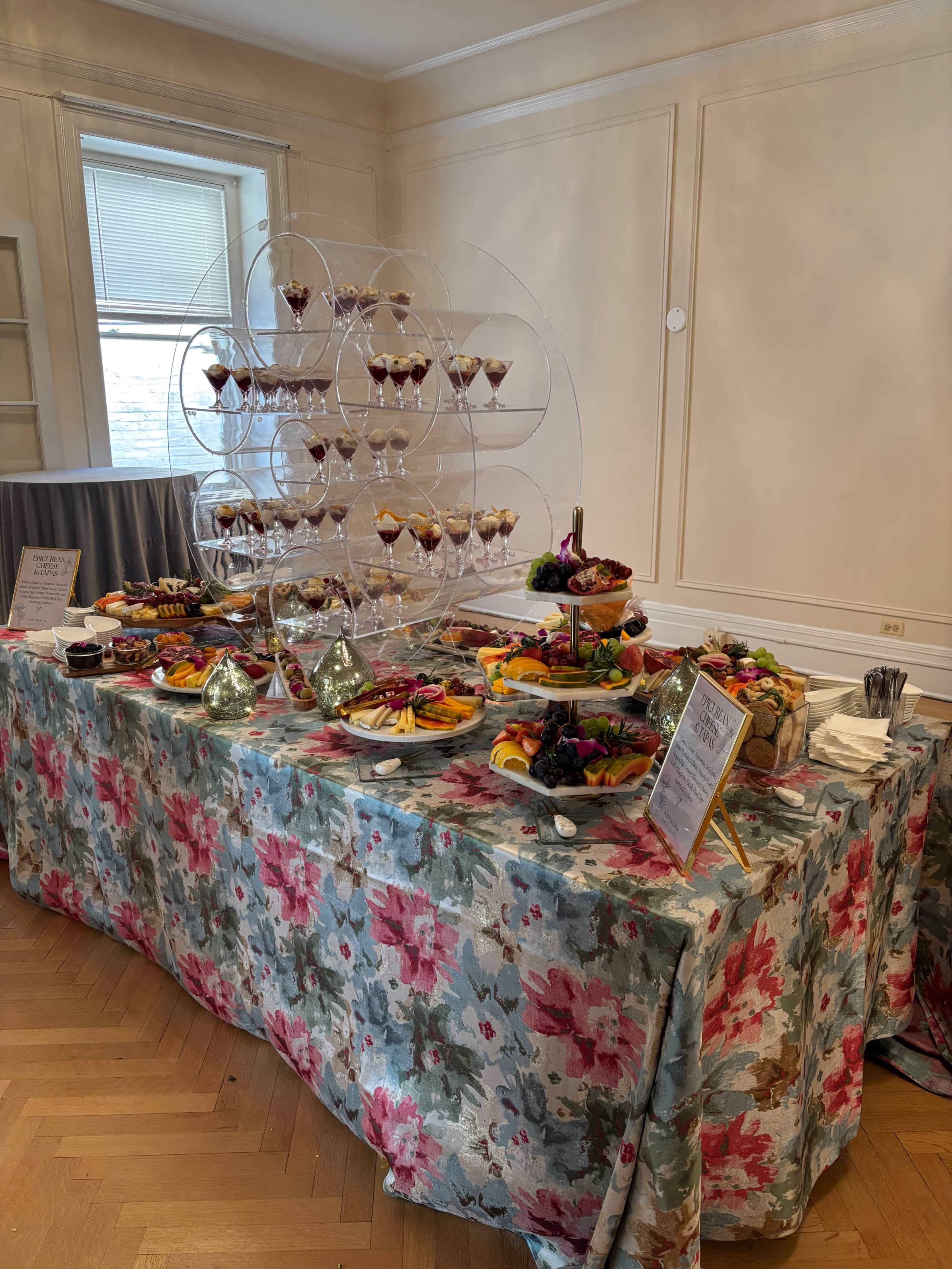 A table is set with a decorative floral tablecloth, featuring an assortment of fruit platters, desserts in glass cups arranged in a tiered stand, and decorative serving utensils.