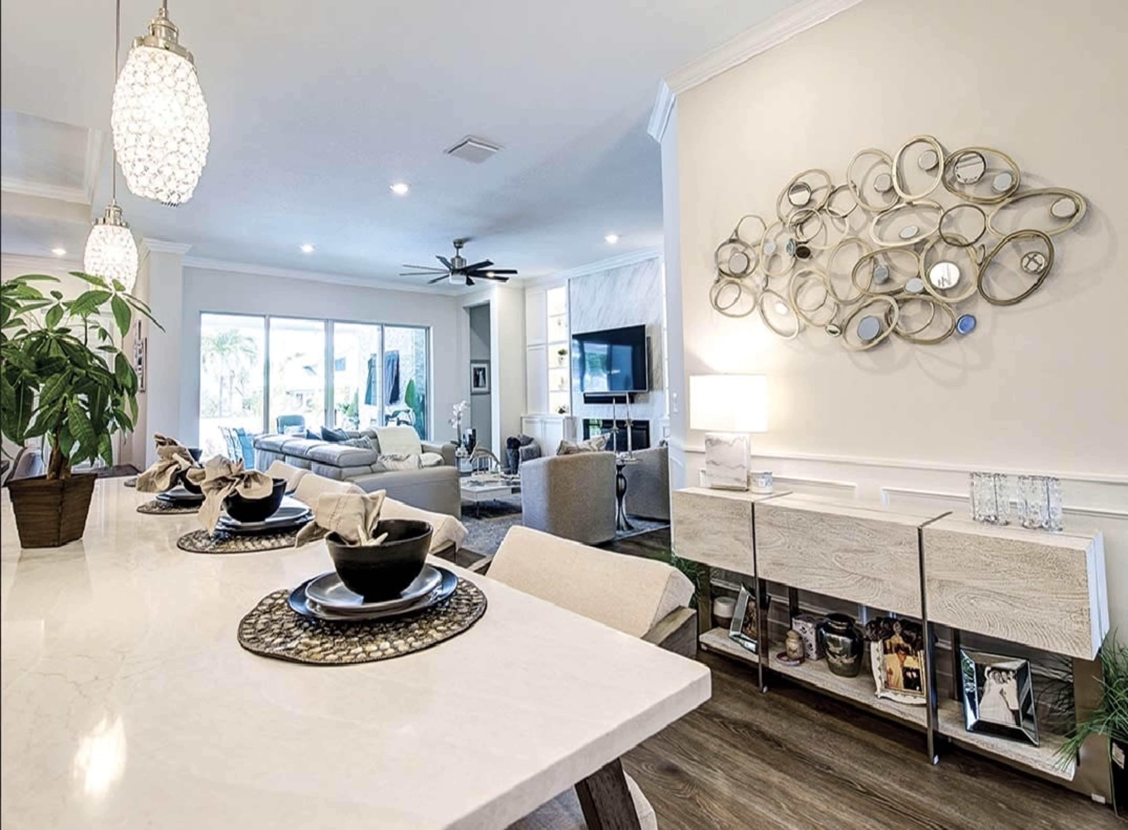 A modern living area features a beige wall with decorative mirrors, a light-colored dining table set with dishes, and a seating area visible in the background with a television.
