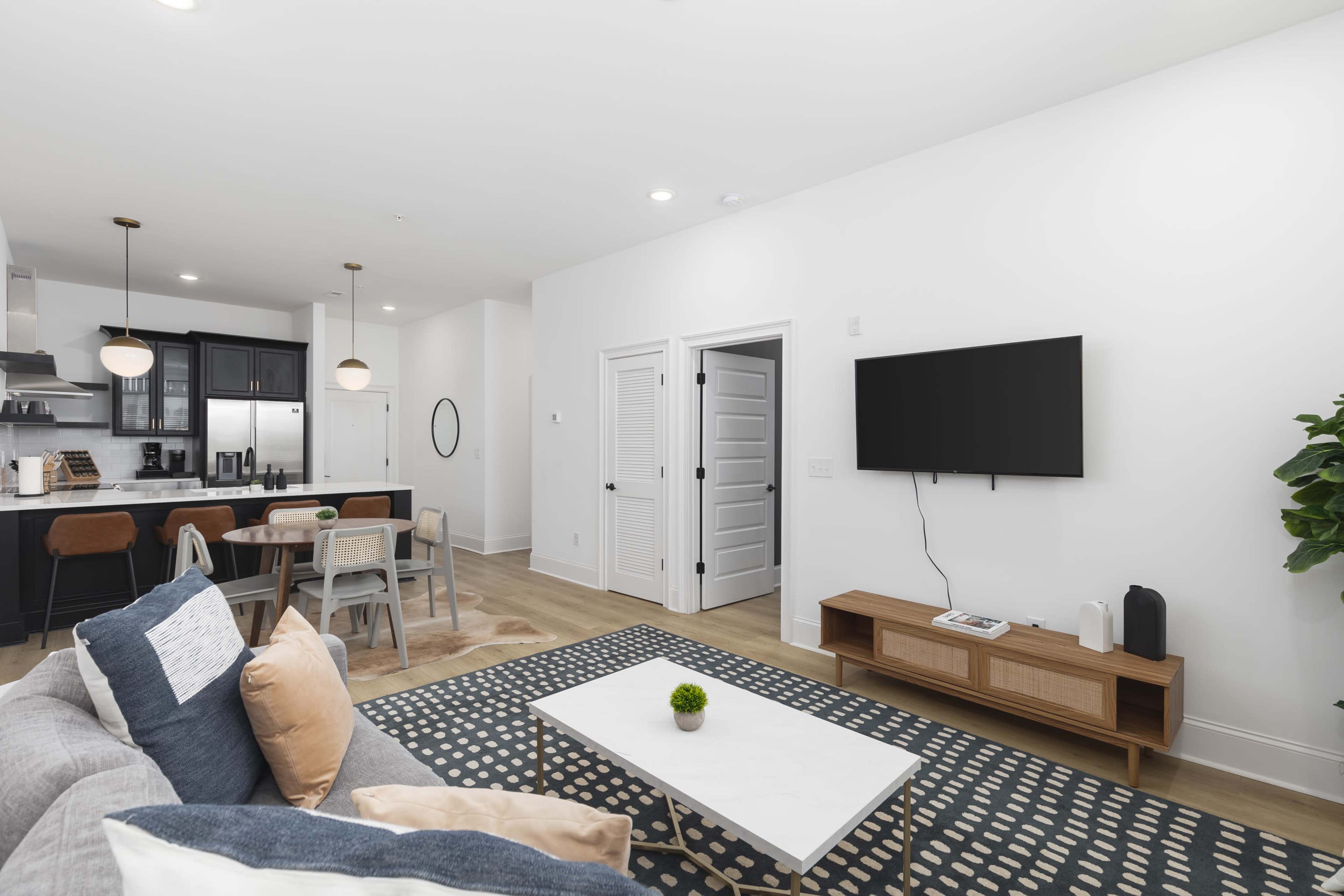 The image shows a modern living room with a gray sofa, a white coffee table, and a wall-mounted television, leading to a dining area with a table and chairs.