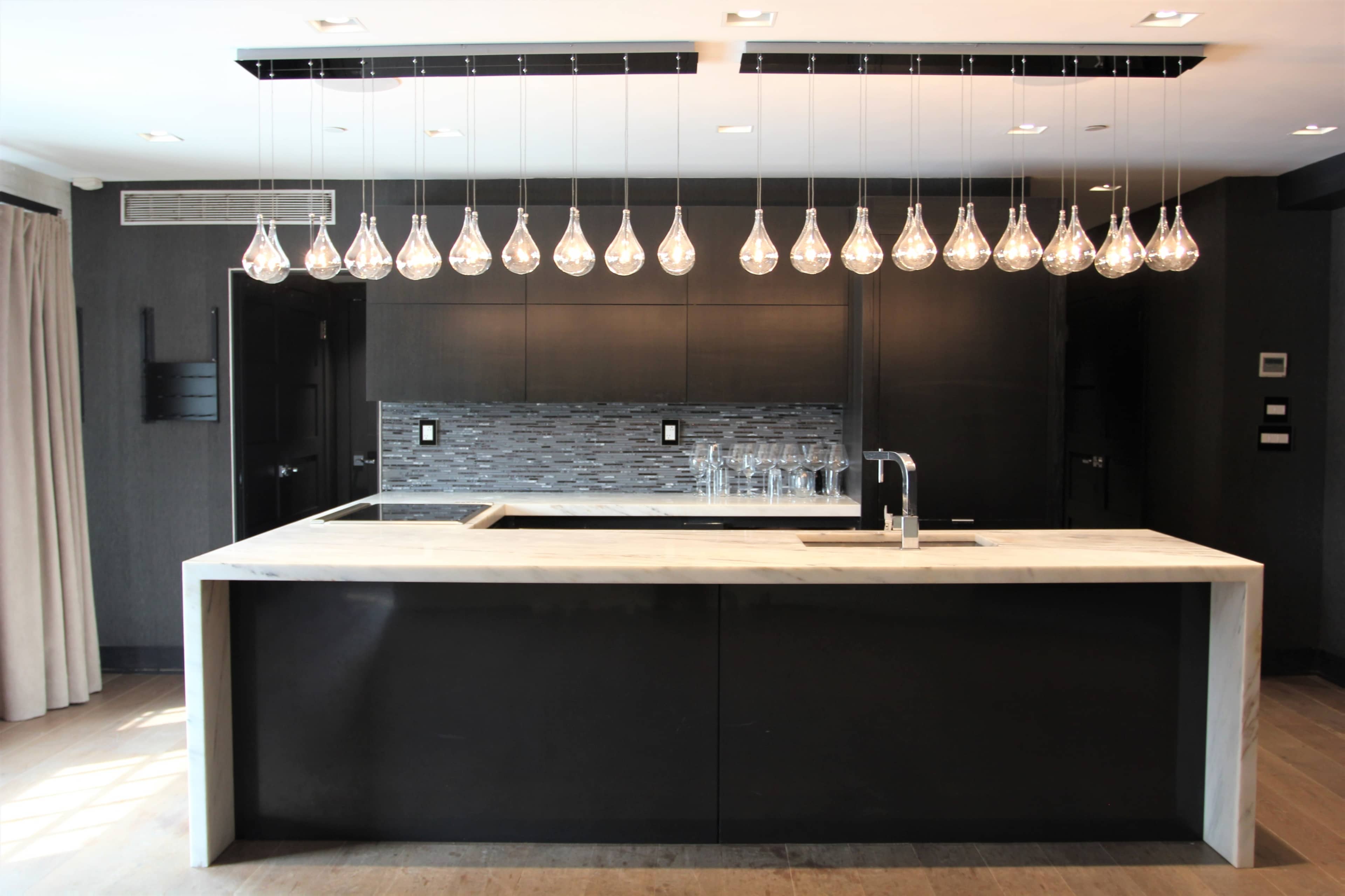 A modern kitchen with a marble island, black cabinetry, and a row of pendant lights hanging above.