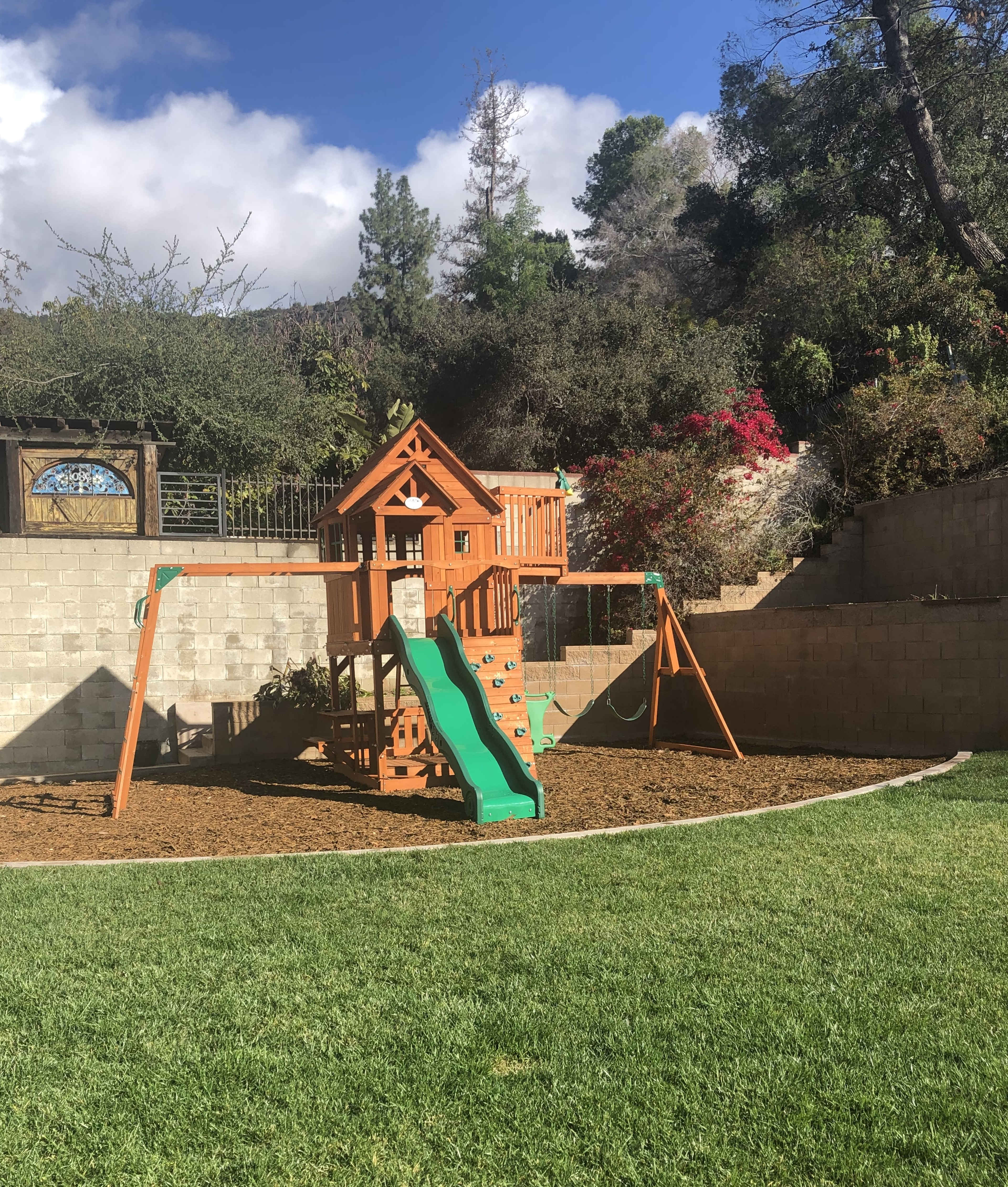 A wooden playground structure with a slide and swings is situated on a grassy area surrounded by a stone wall and greenery.