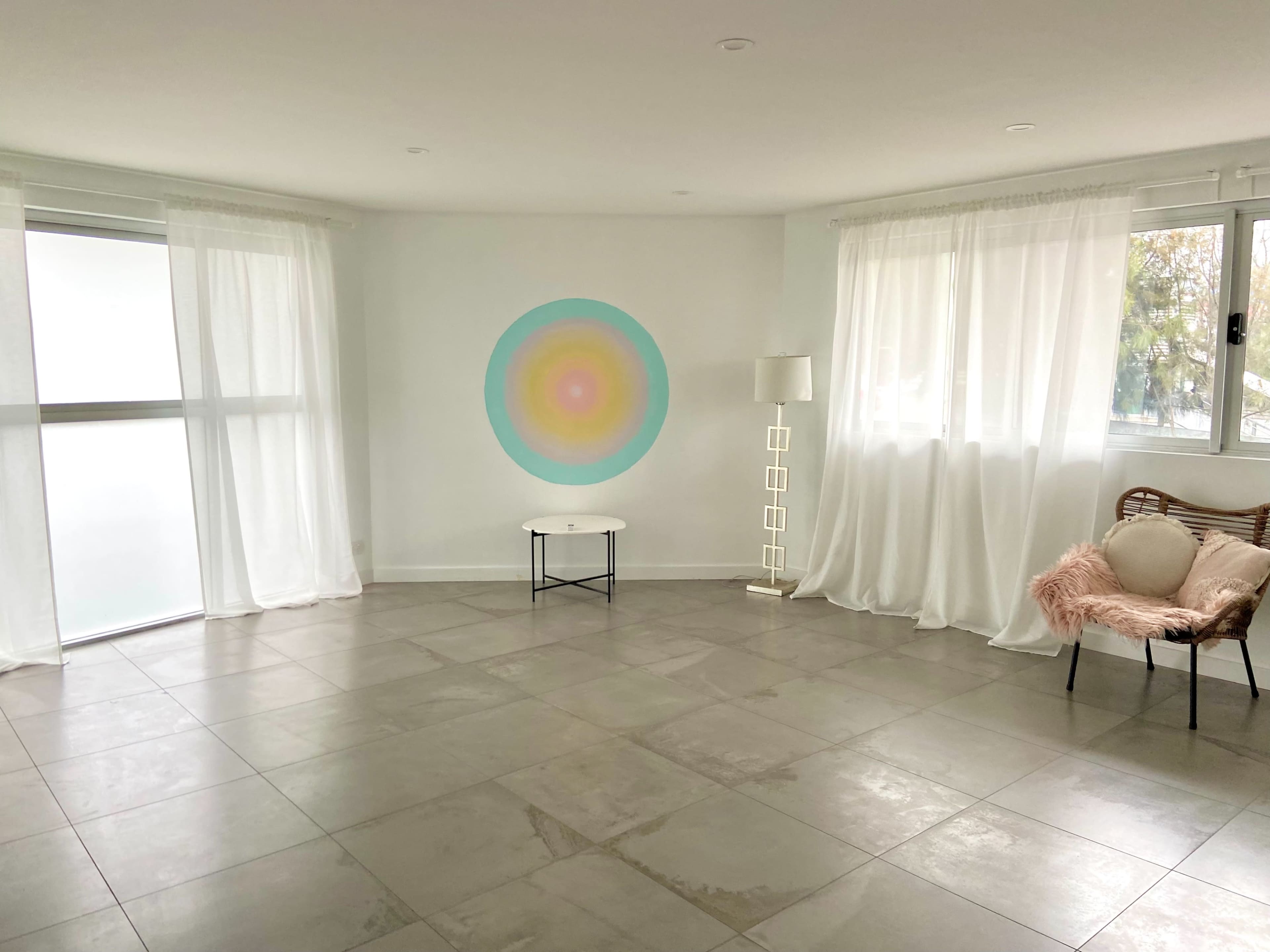 A spacious room features large windows with sheer curtains, a circular wall design in pastel colors, a small black coffee table, and a patterned armchair with a throw.