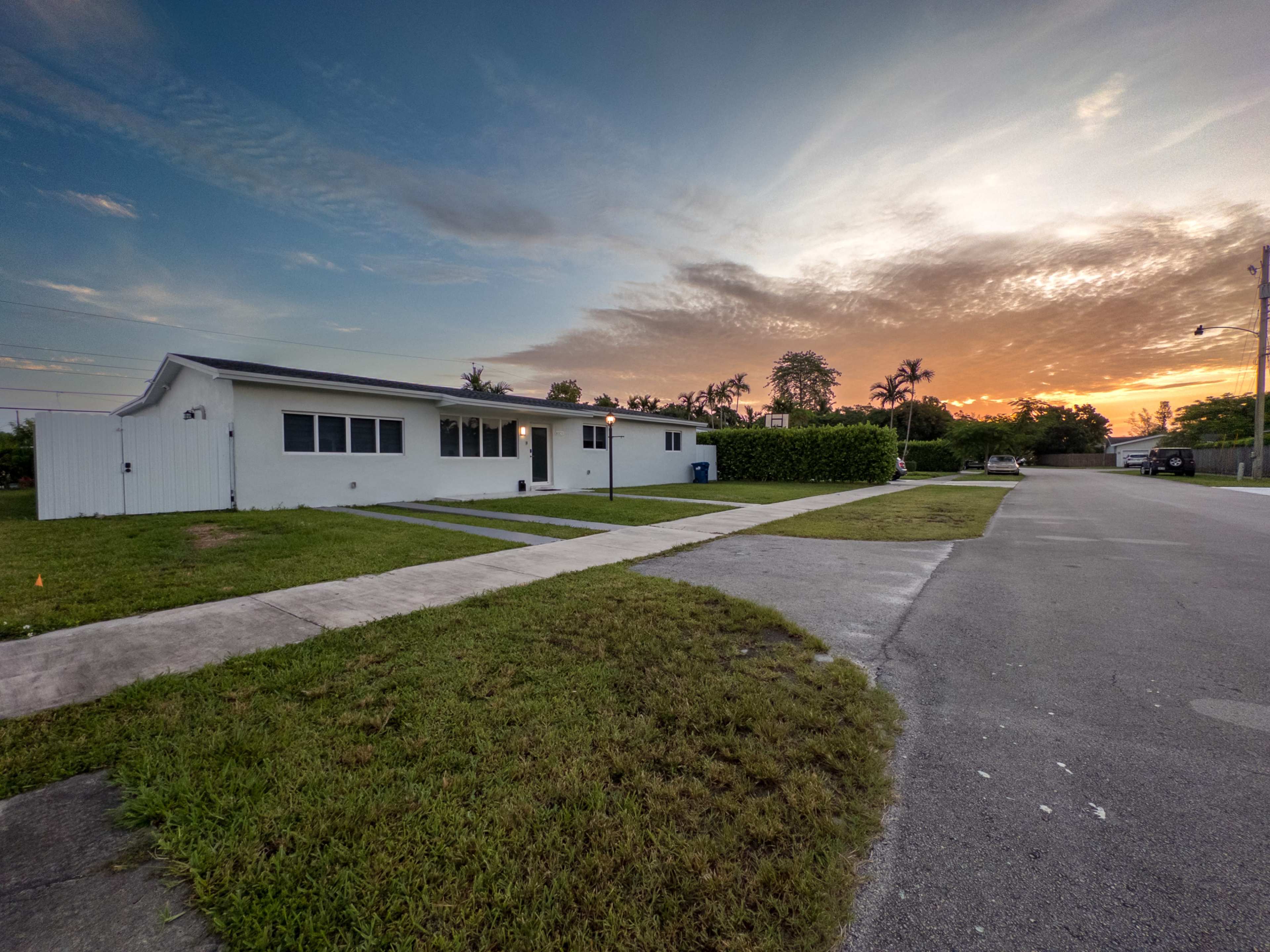 A single-story white house sits on a corner lot with a driveway, under a colorful sunset sky.