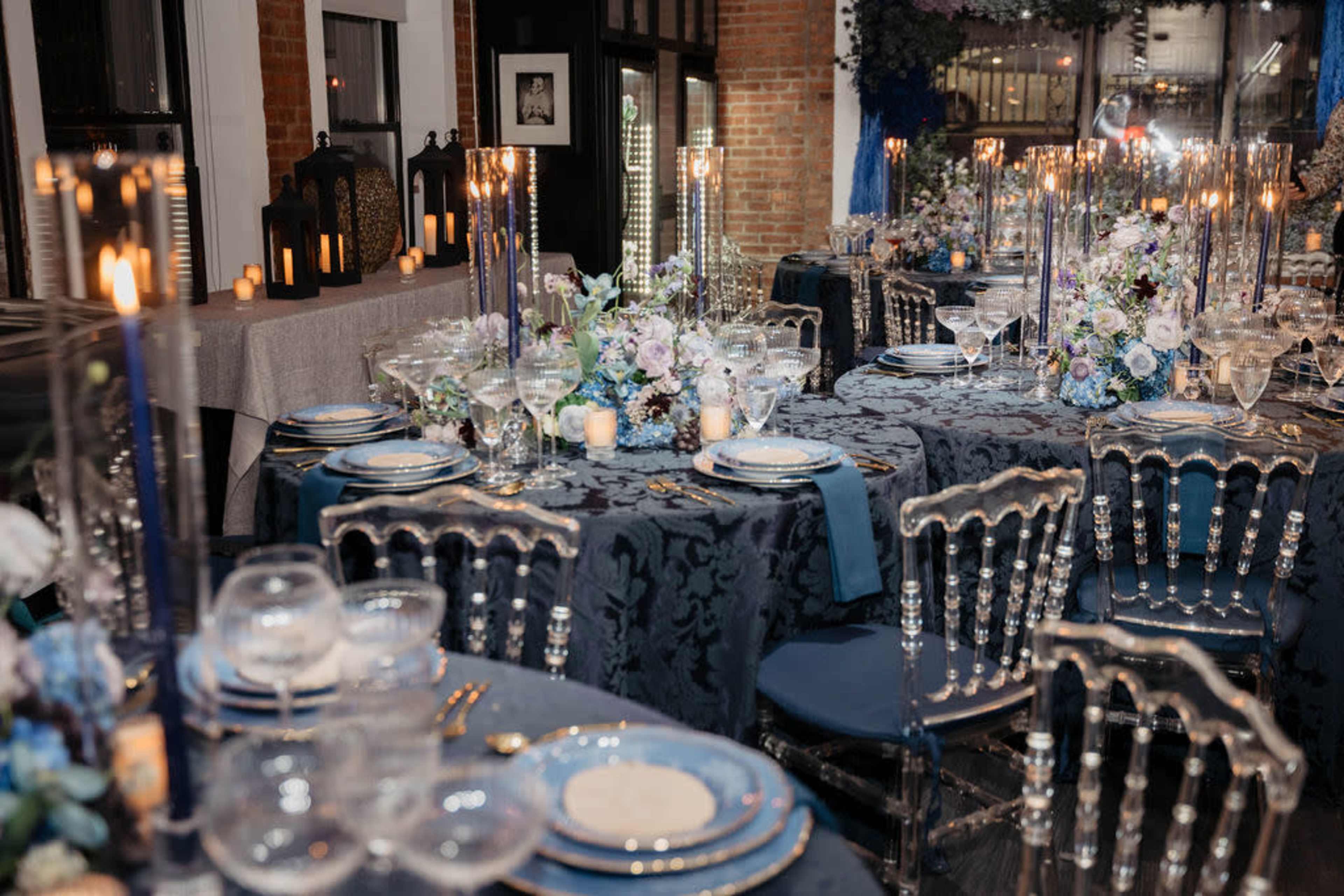 The image shows a beautifully arranged banquet hall with elegantly set tables featuring dark blue tablecloths, fine china, crystal glassware, and tall candlesticks, surrounded by floral centerpieces.