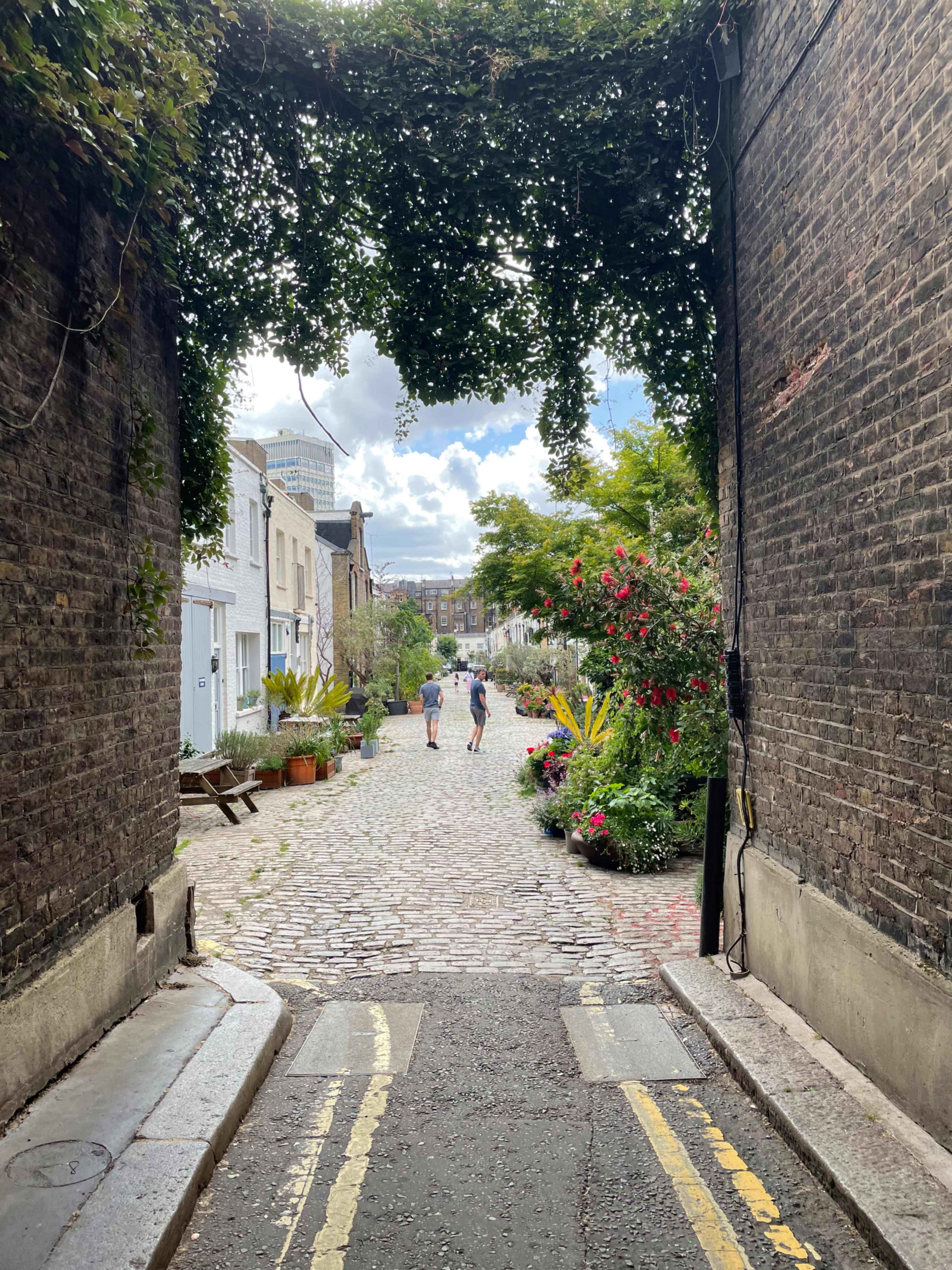 A cobblestone alley lined with greenery and colorful flowers leads to a street where two people walk toward the camera.