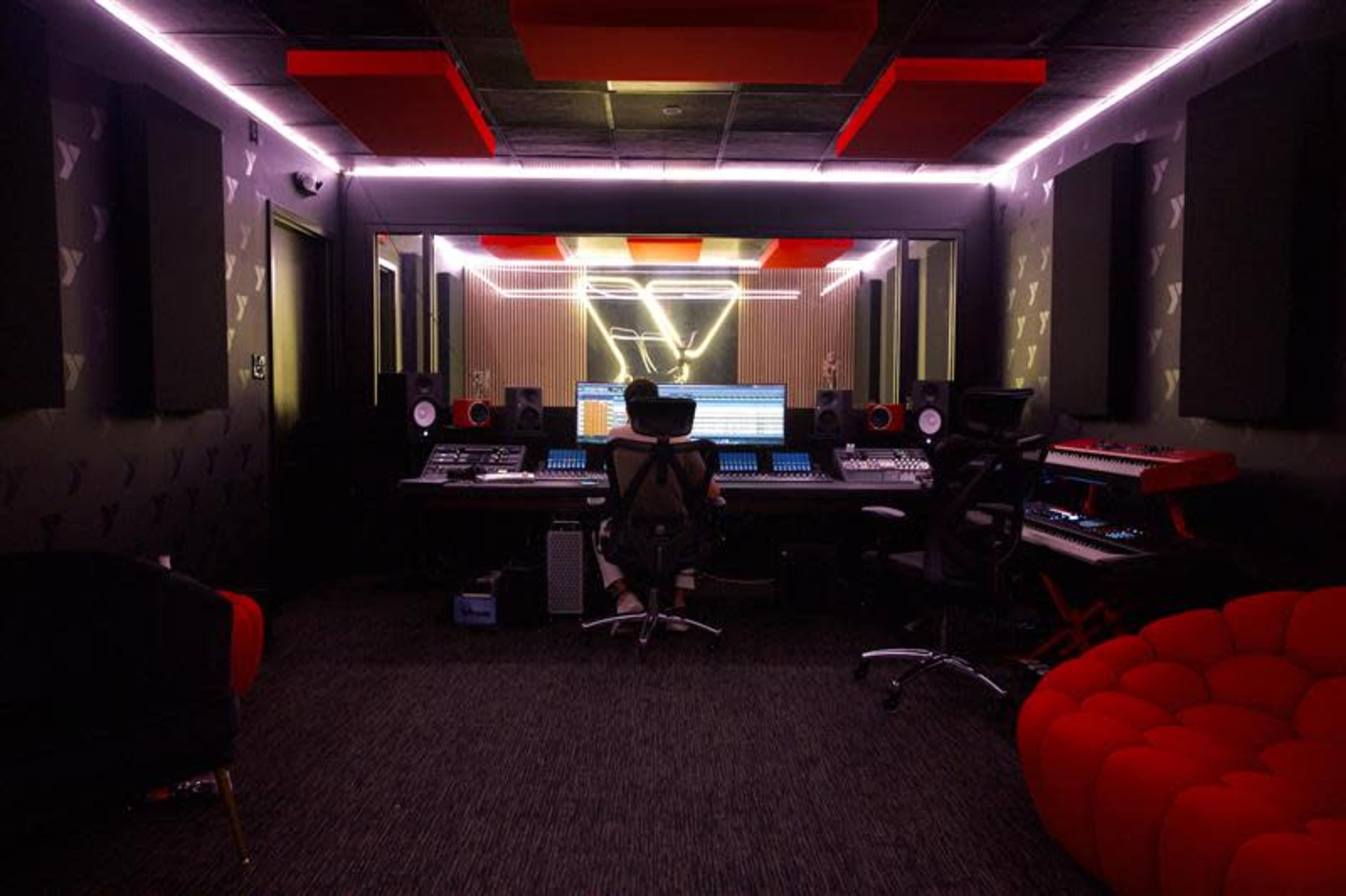 A person sits at a large mixing console in a recording studio, surrounded by various audio equipment and illuminated by neon lighting.