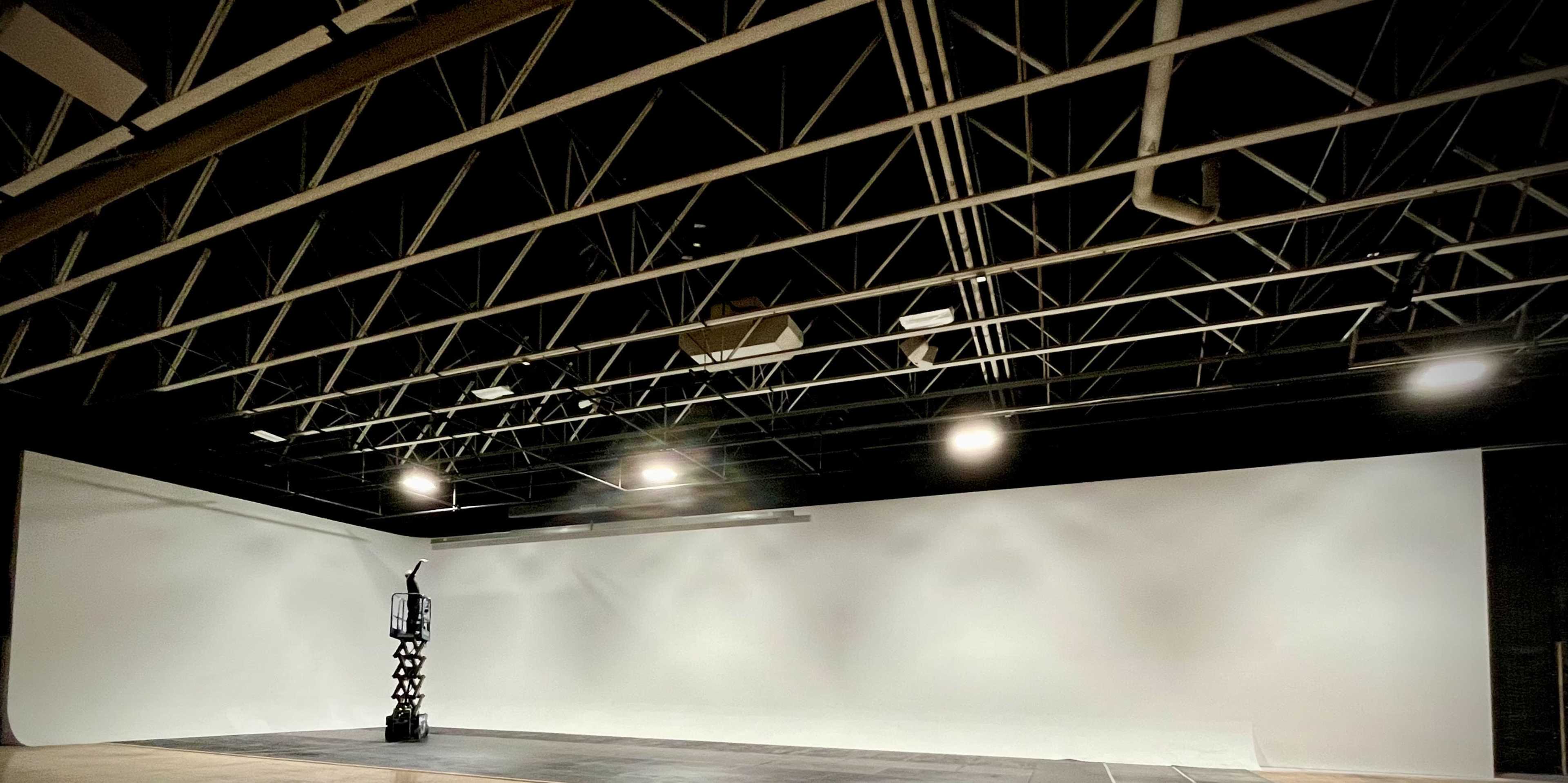 The image shows an empty studio space with a white backdrop, exposed beams, and overhead lighting.