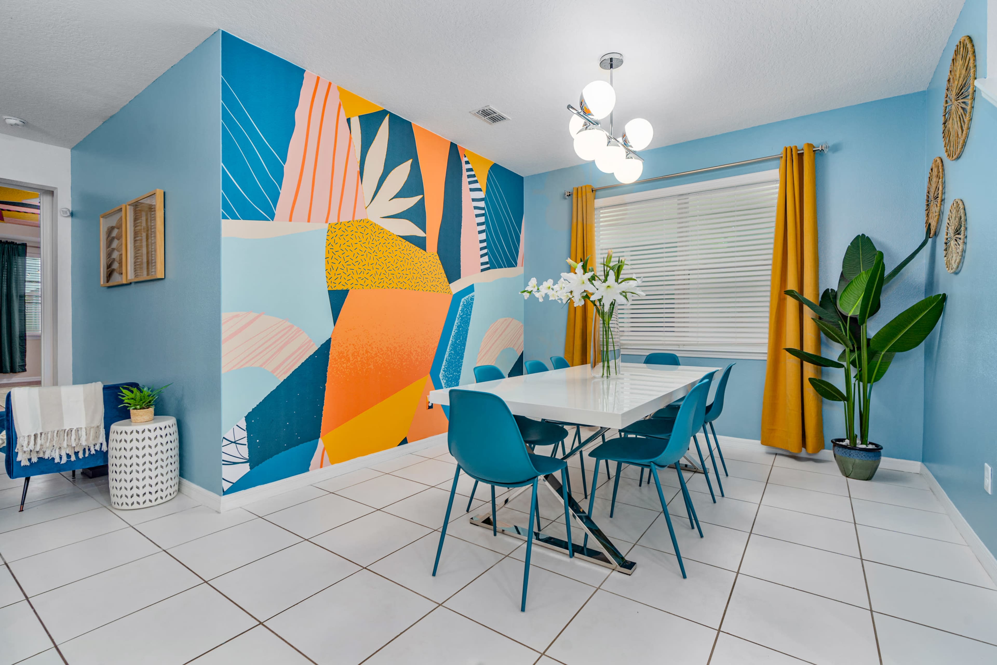A modern dining area features a large white table surrounded by blue chairs, against a vibrant mural and bright yellow curtains.
