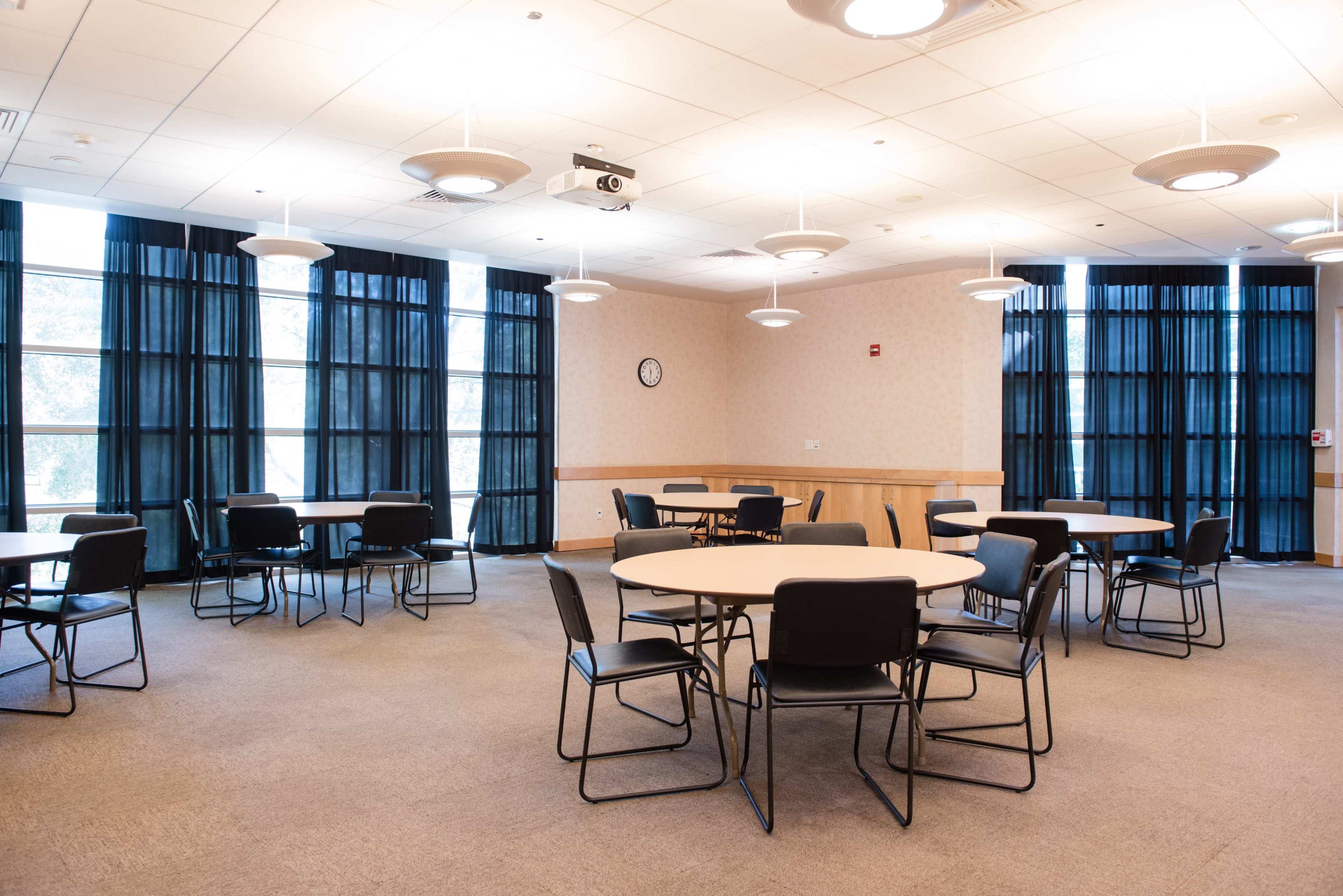 A room with several round tables and black chairs arranged for a meeting, featuring large windows covered with black curtains.