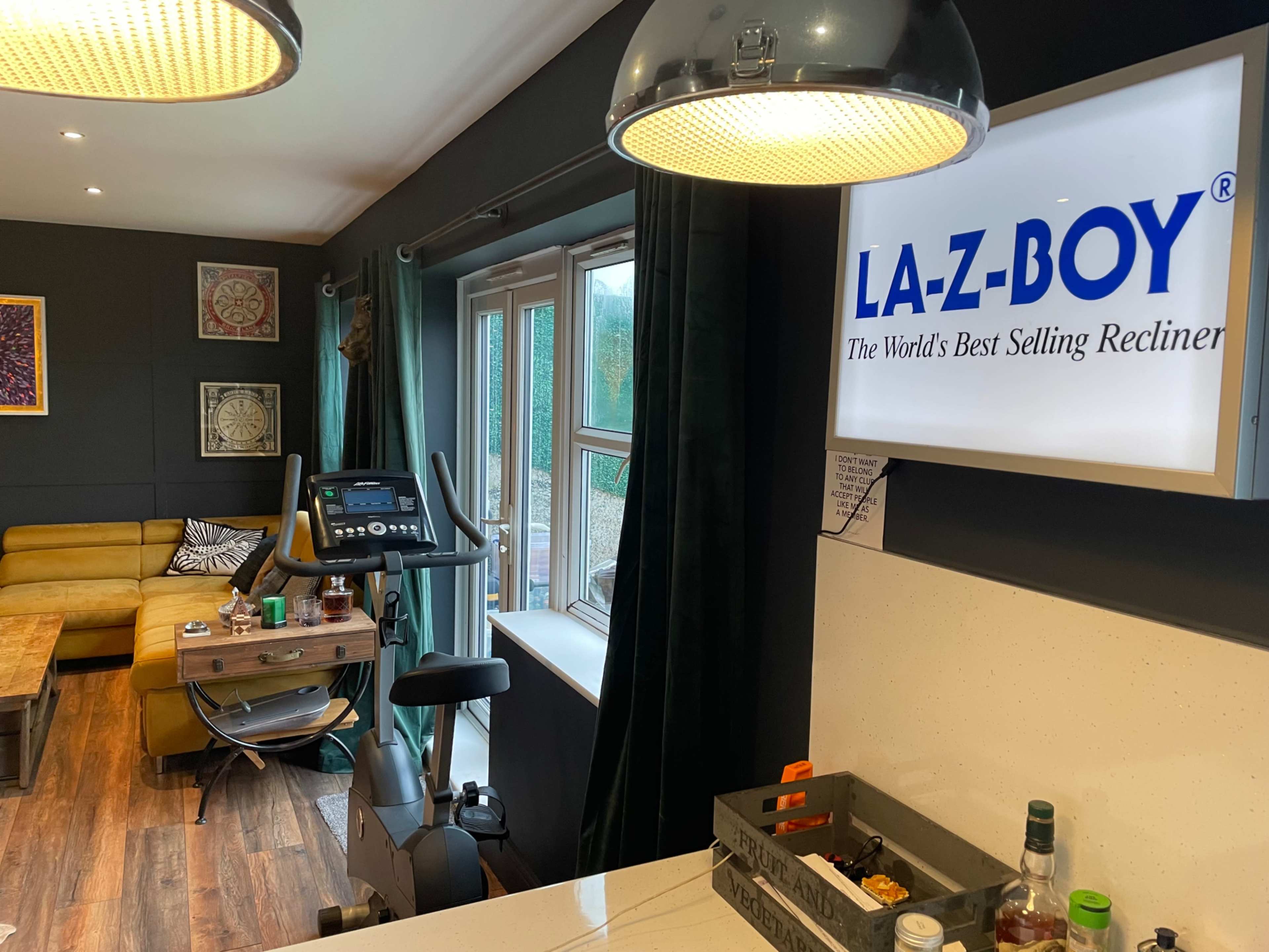 The image shows a living space featuring a yellow sofa, a wooden coffee table, an exercise bike, and a prominently displayed La-Z-Boy sign on the wall.