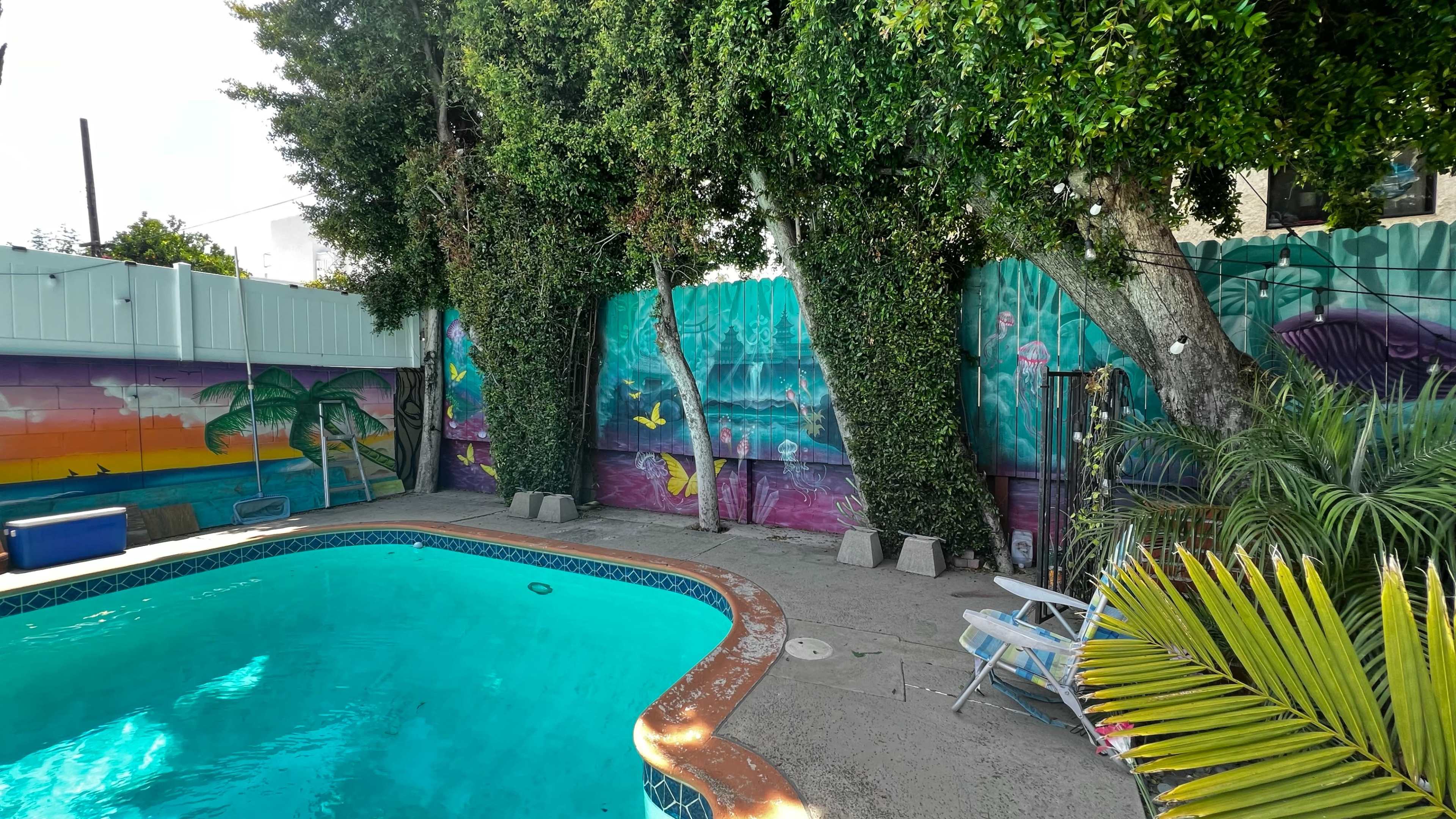 A pool area features vibrant muraled walls, lush greenery, and lawn chairs beside the water.