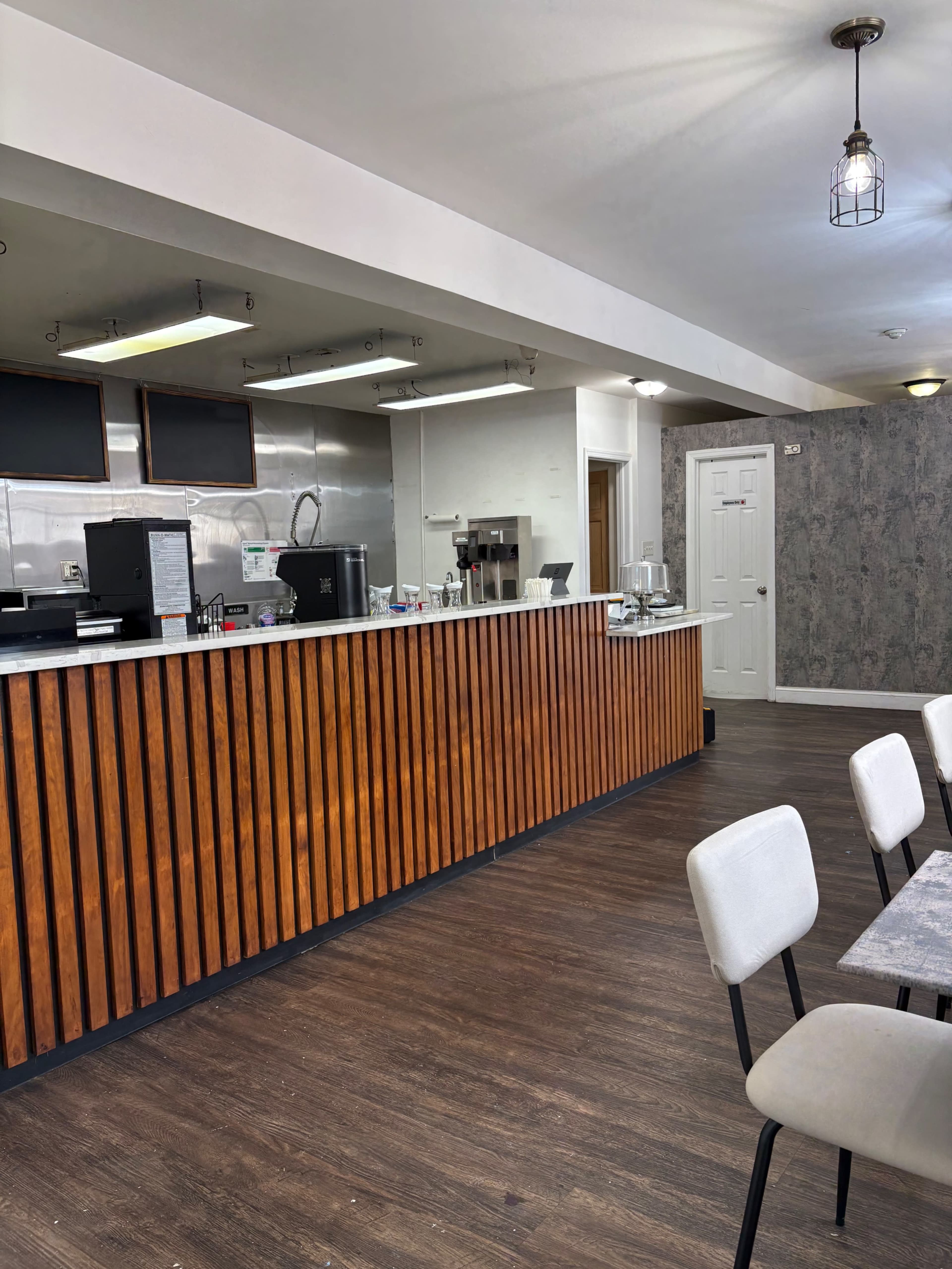 The image shows a modern café interior with a wooden counter, stainless steel behind it, and a seating area featuring light-colored chairs and a patterned wall.