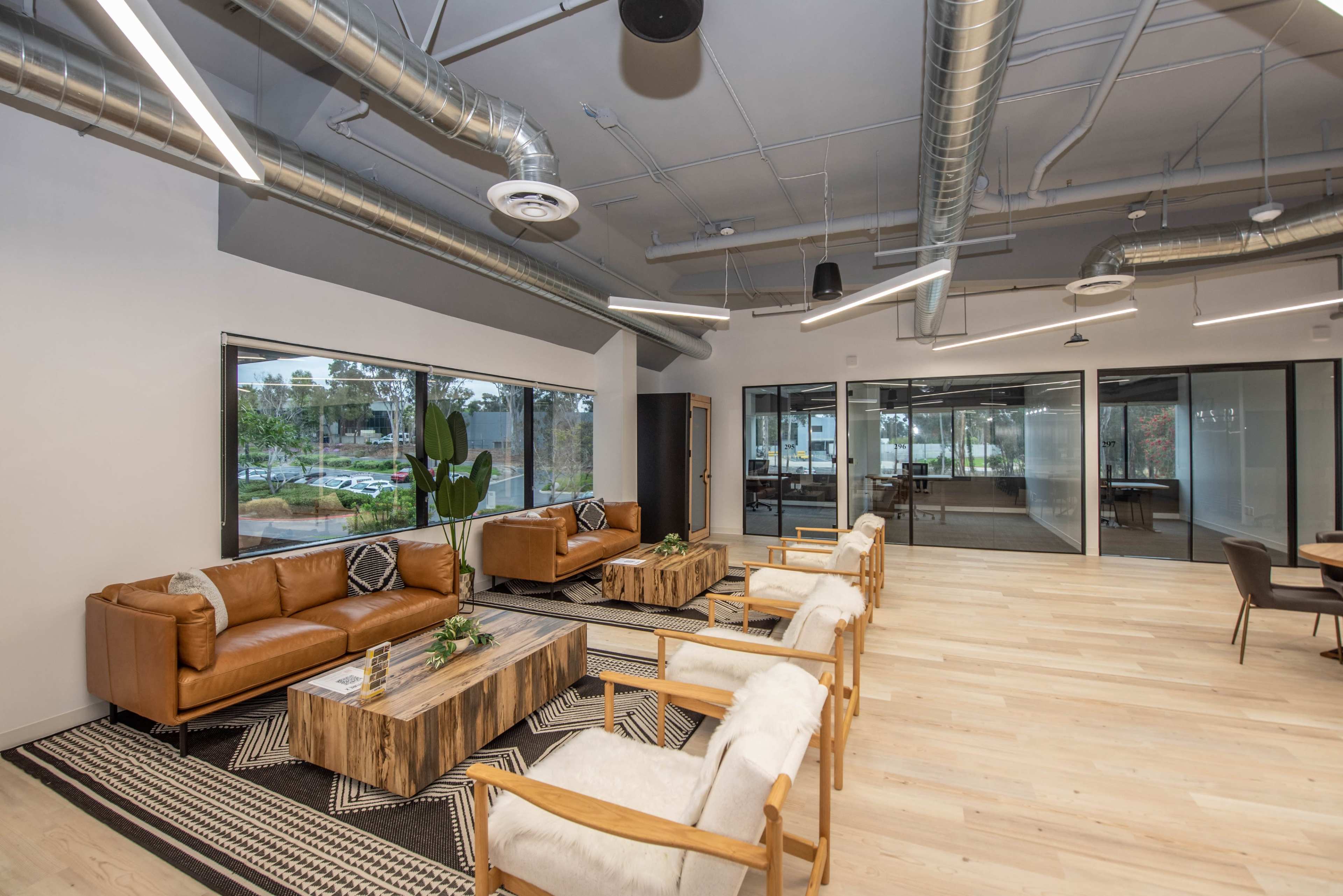 A modern office lounge area with leather sofas, wooden coffee tables, and a large window overlooking a green landscape.