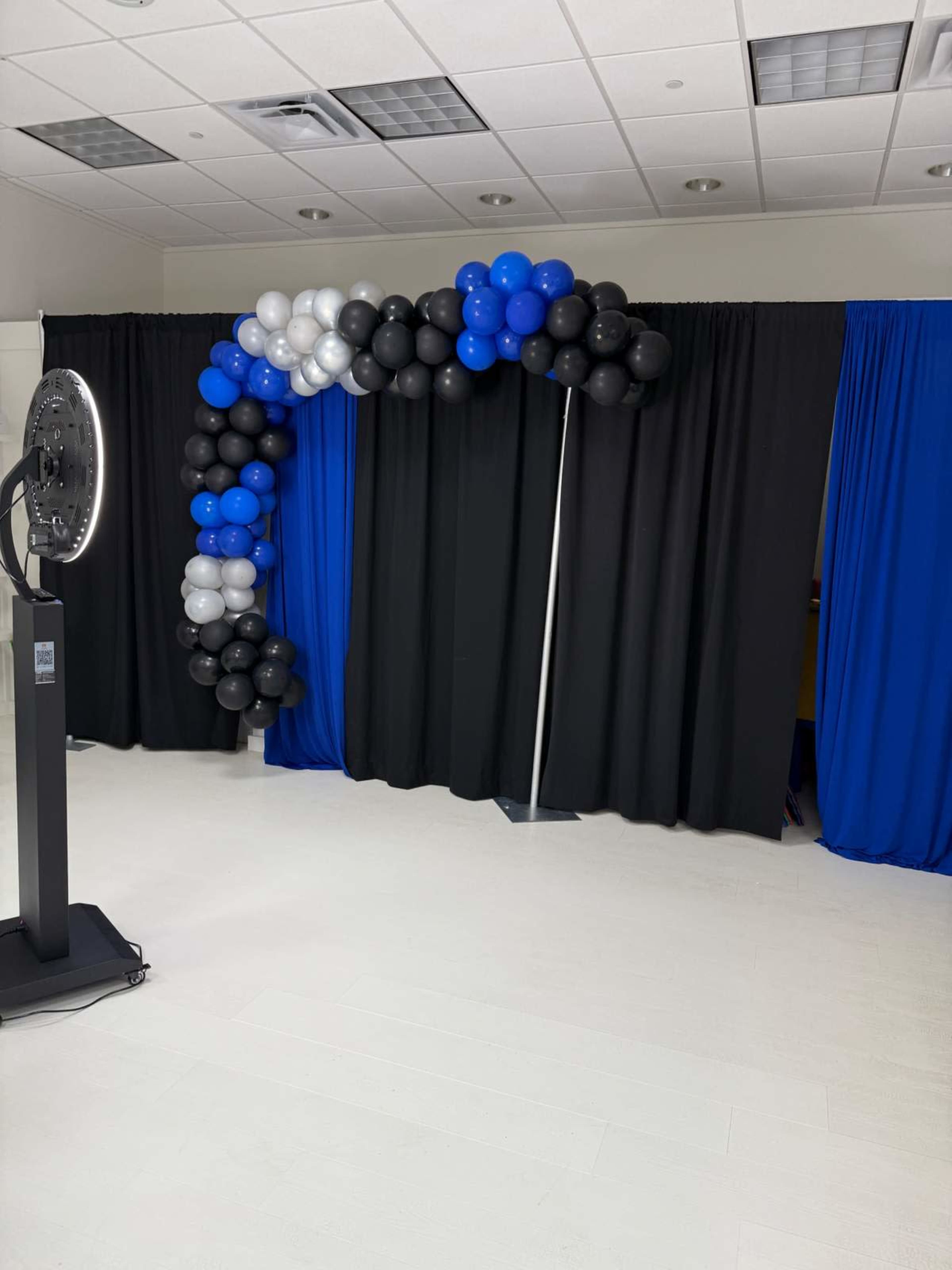 The image shows an arch made of blue, black, and silver balloons, next to black and blue fabric drapes in a well-lit indoor space.