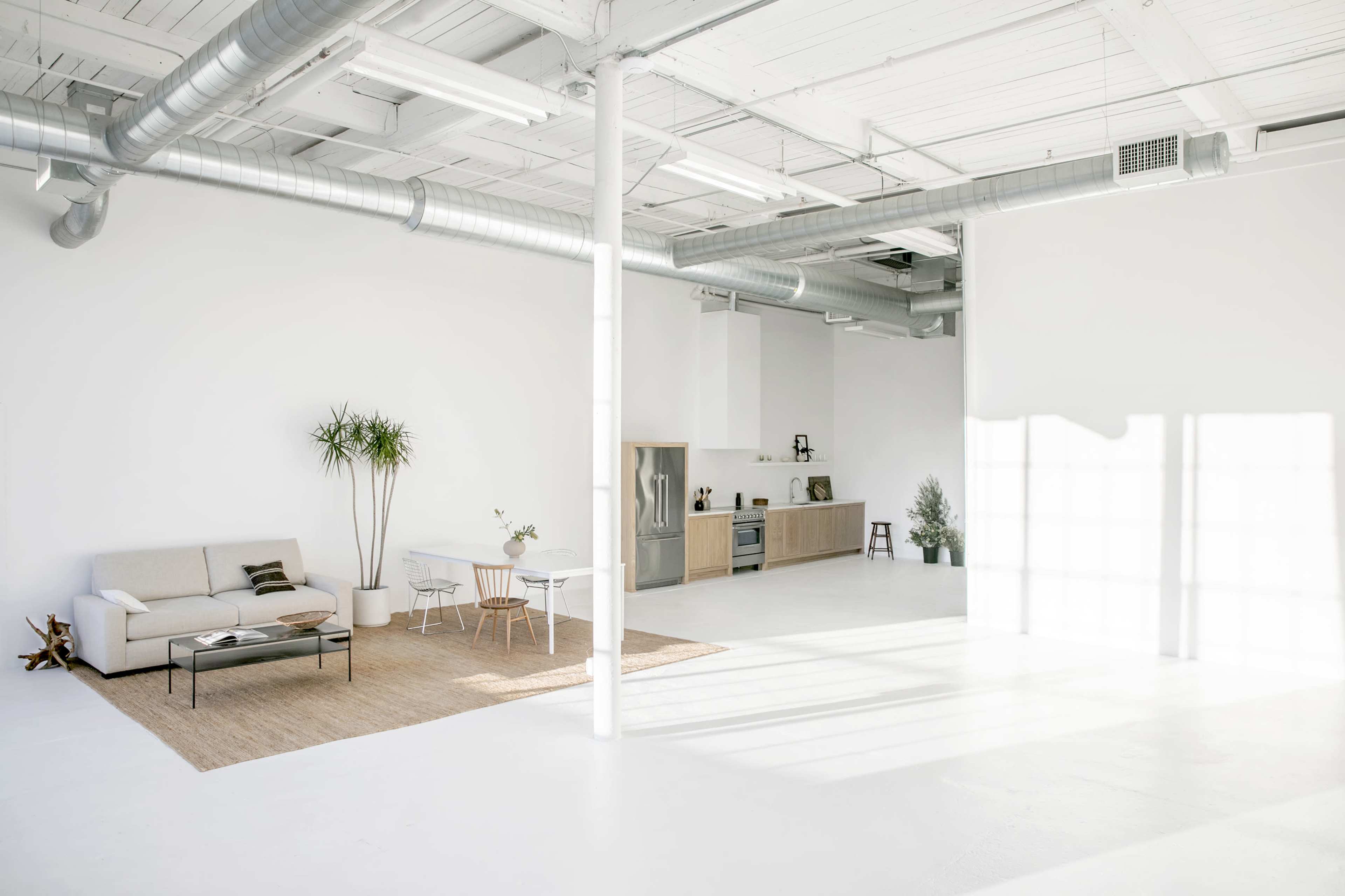 The image displays a minimalist interior with a sofa, dining table, and kitchen area under bright natural light.