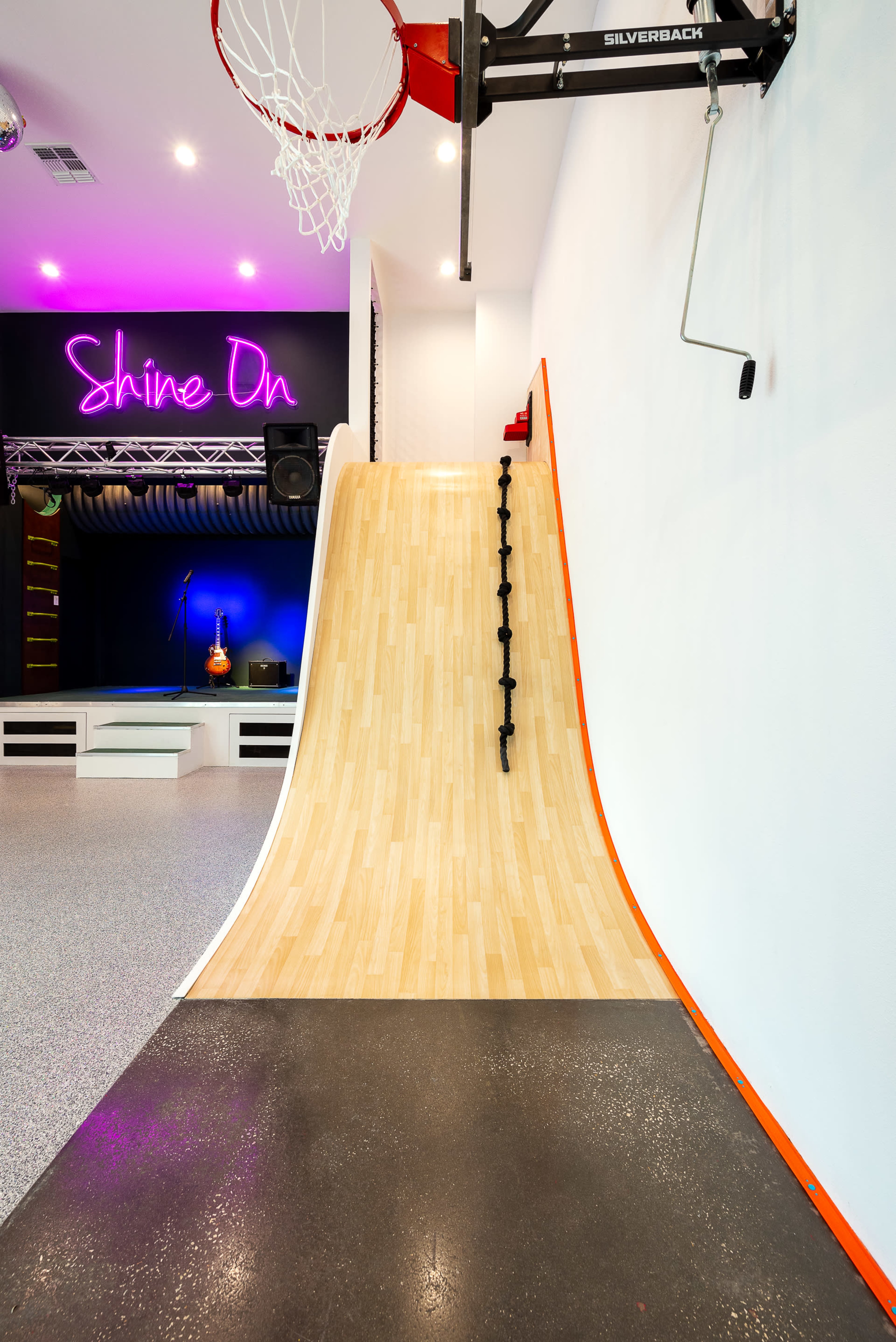 The image shows a basketball hoop next to a smooth wooden ramp with a black grip for climbing, set against a brightly lit wall with neon text.