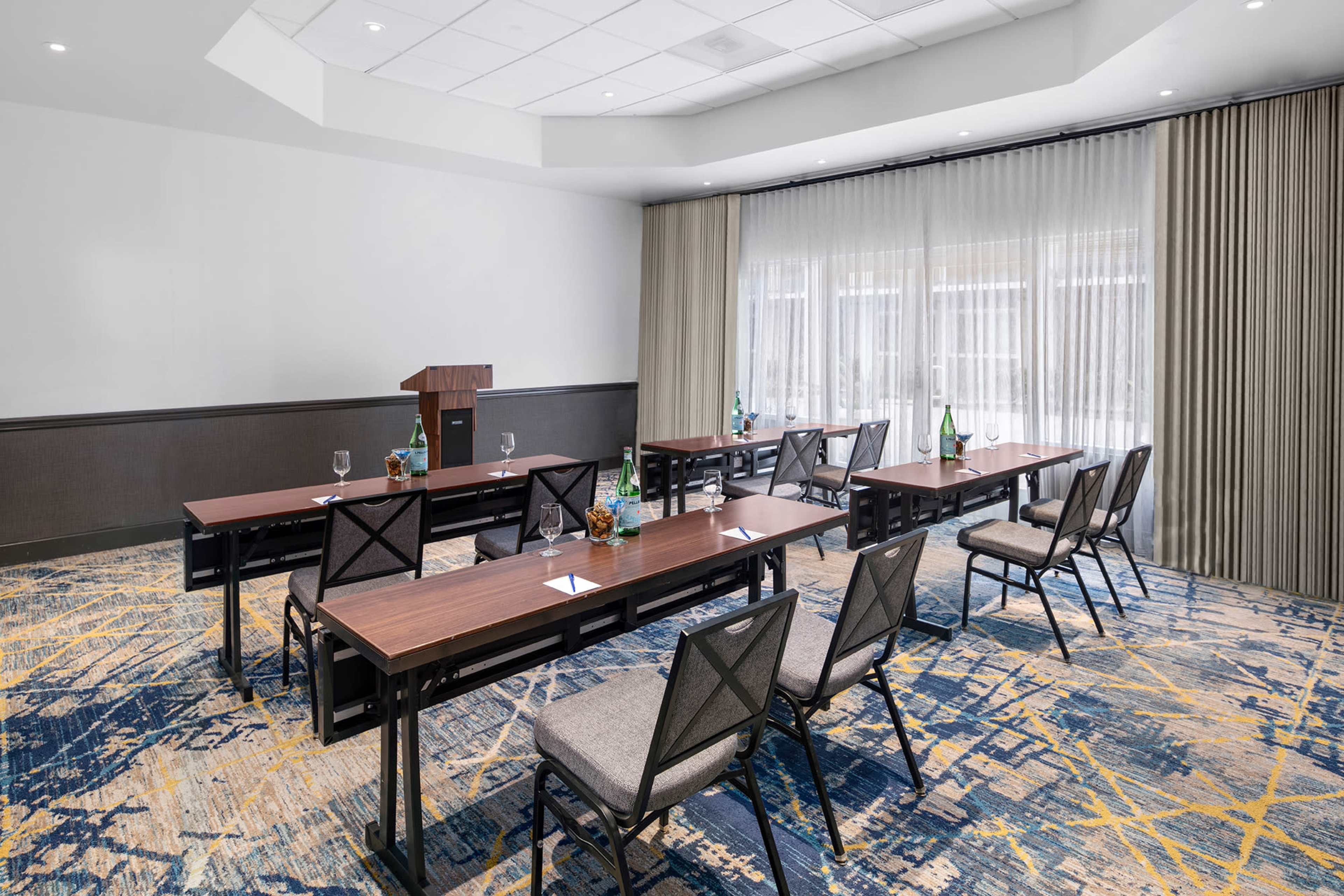 A conference room features several rows of tables with chairs arranged for a meeting, along with a podium and natural light coming through large windows.
