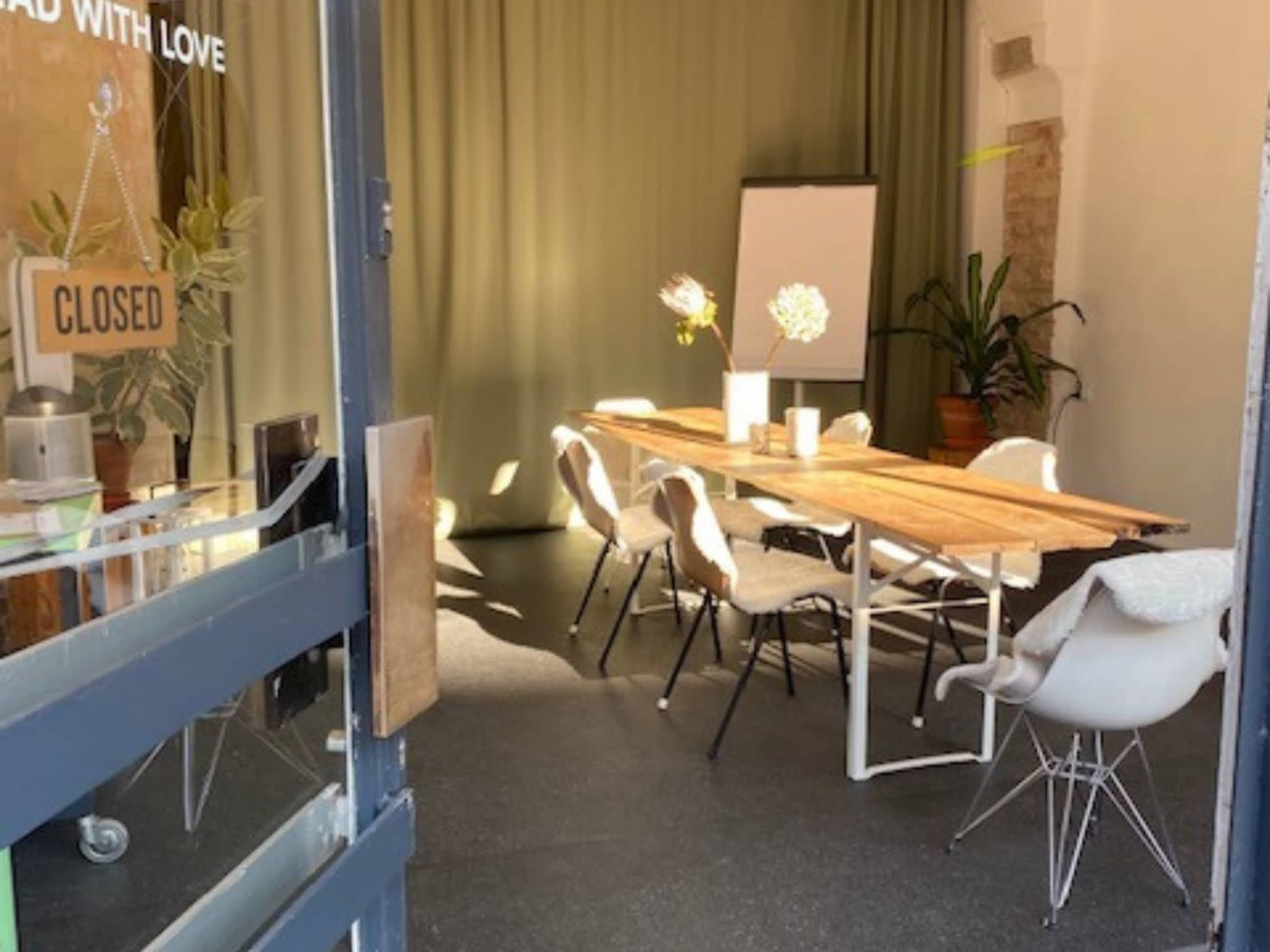 A conference room with a long wooden table and white chairs is set up under soft lighting, featuring a blank flip chart and potted plants in the background.