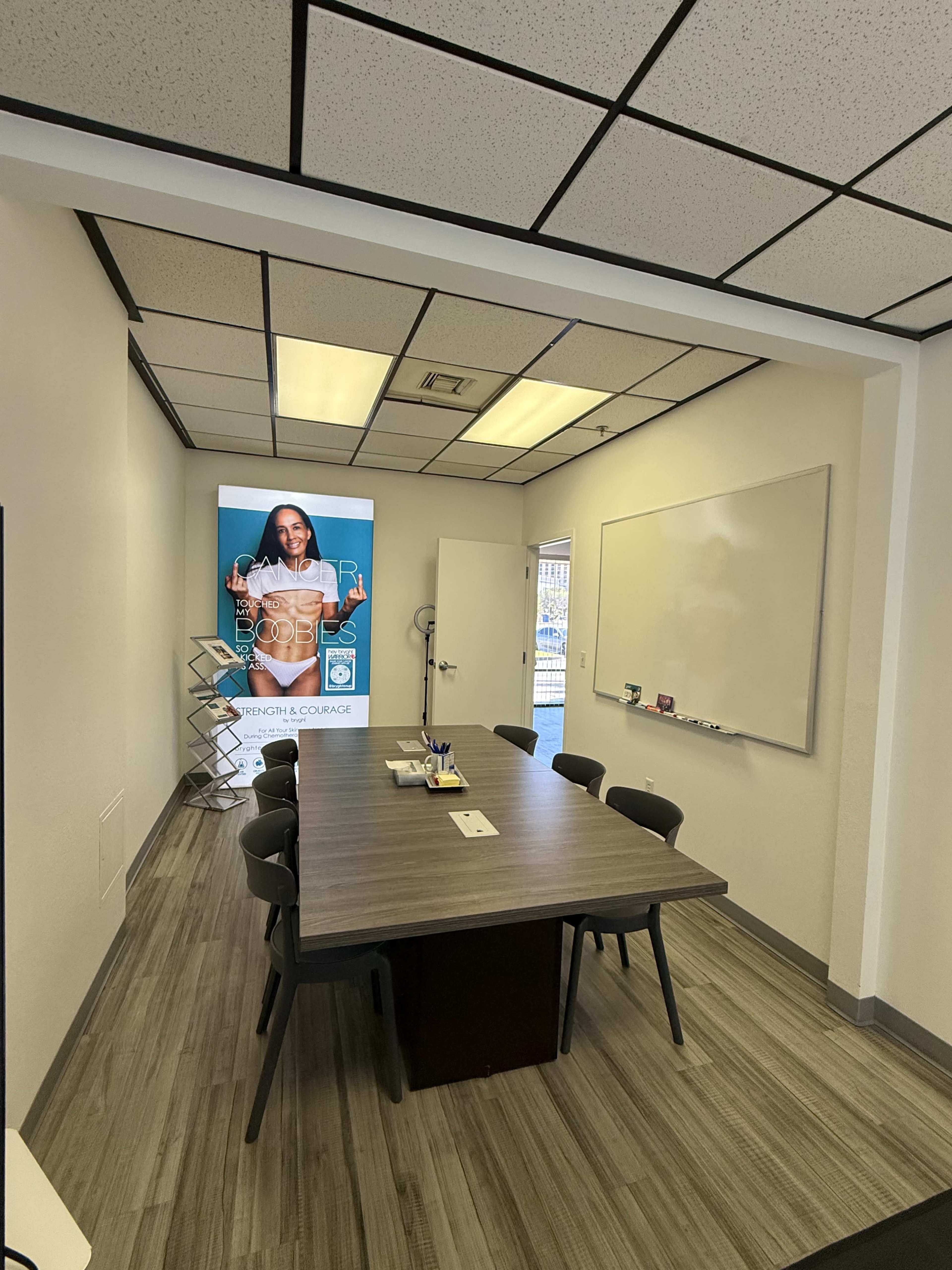 The image shows a conference room with a long table, chairs, a whiteboard, and a promotional poster on the wall.