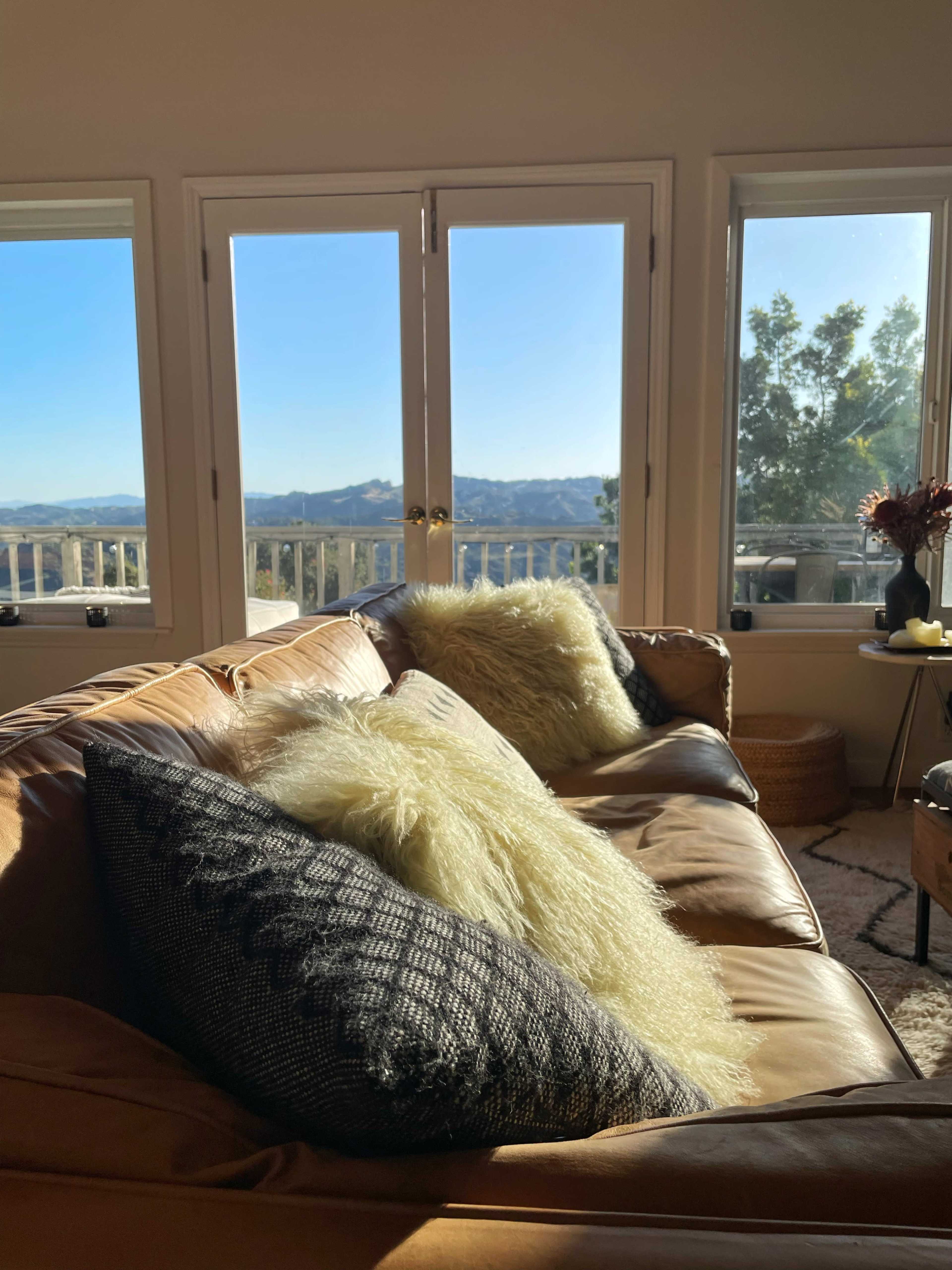 A leather sofa with textured pillows is positioned in front of large windows that offer a view of distant mountains and a clear blue sky.