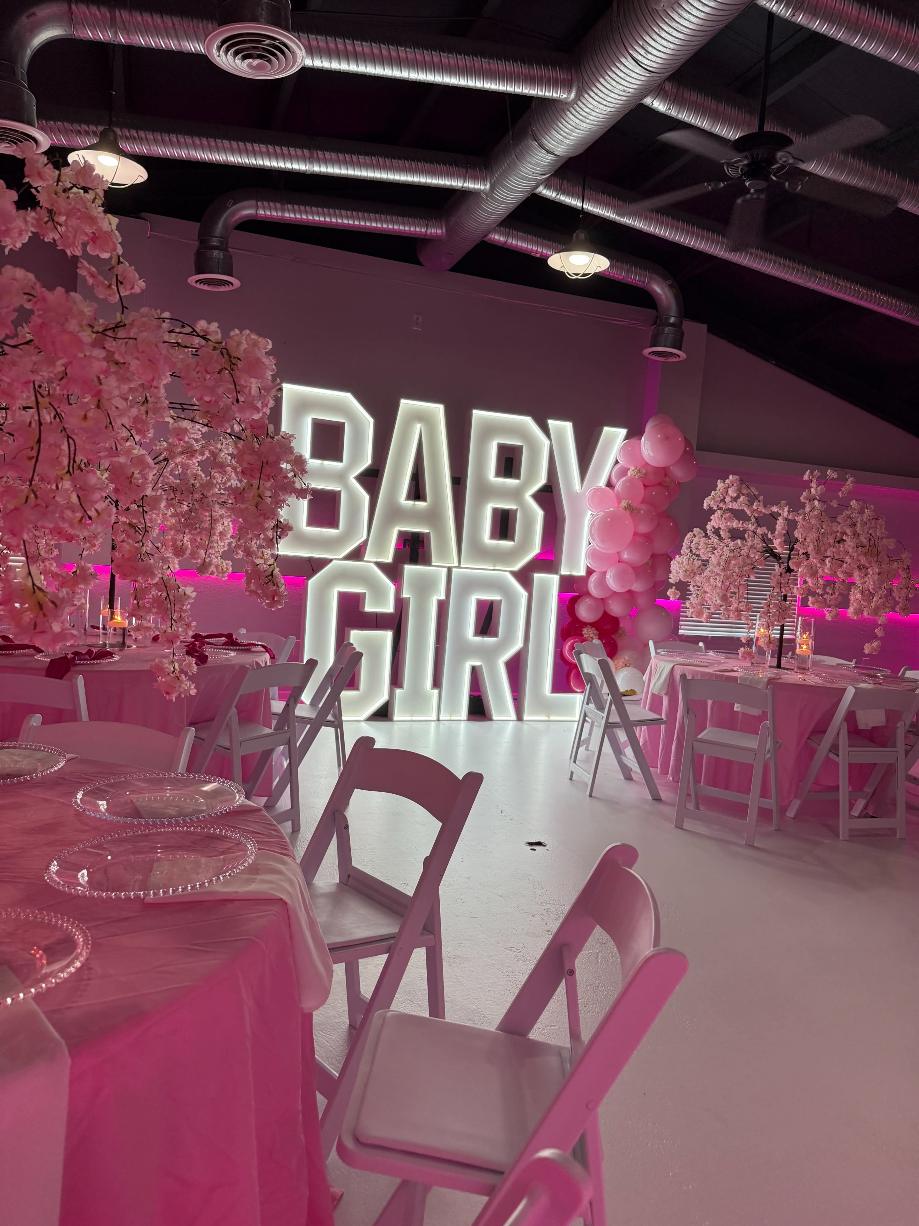 The image shows a decorated indoor space featuring pink flowering trees, pink lighting, and a large illuminated "BABY GIRL" sign, with tables set for an event.