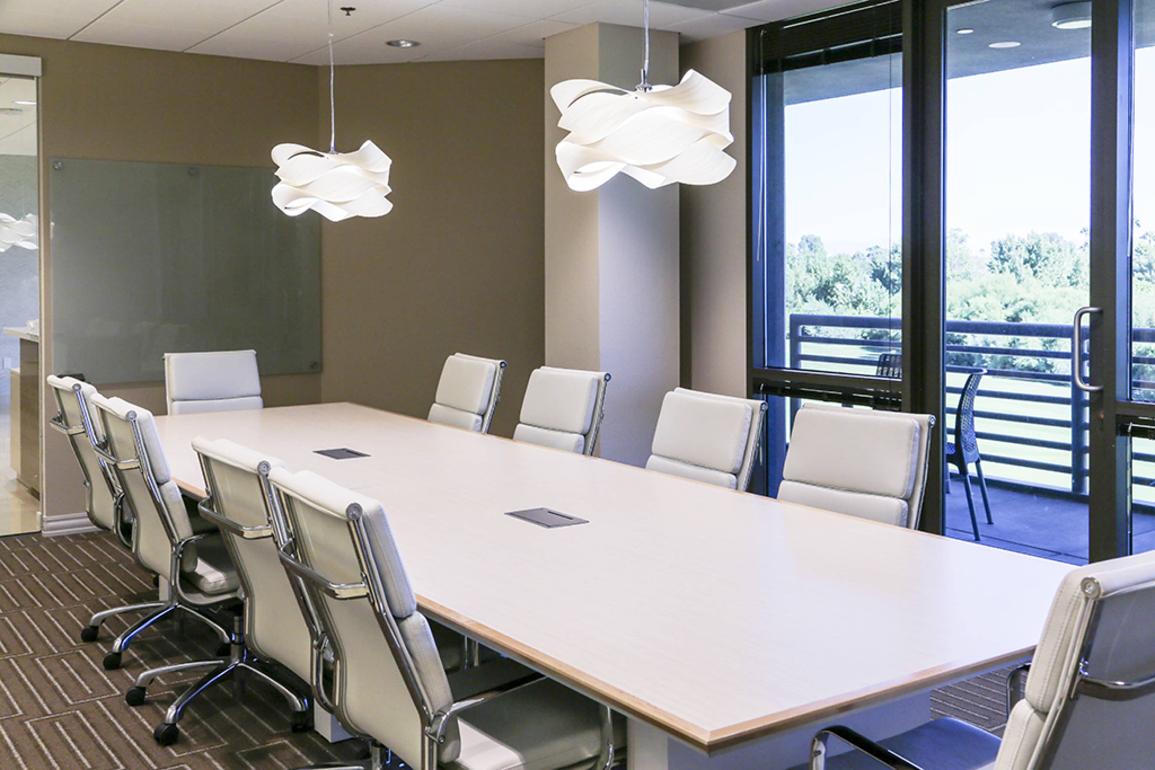 A spacious conference room features a long wooden table surrounded by modern white chairs, with large windows providing views of greenery outside.
