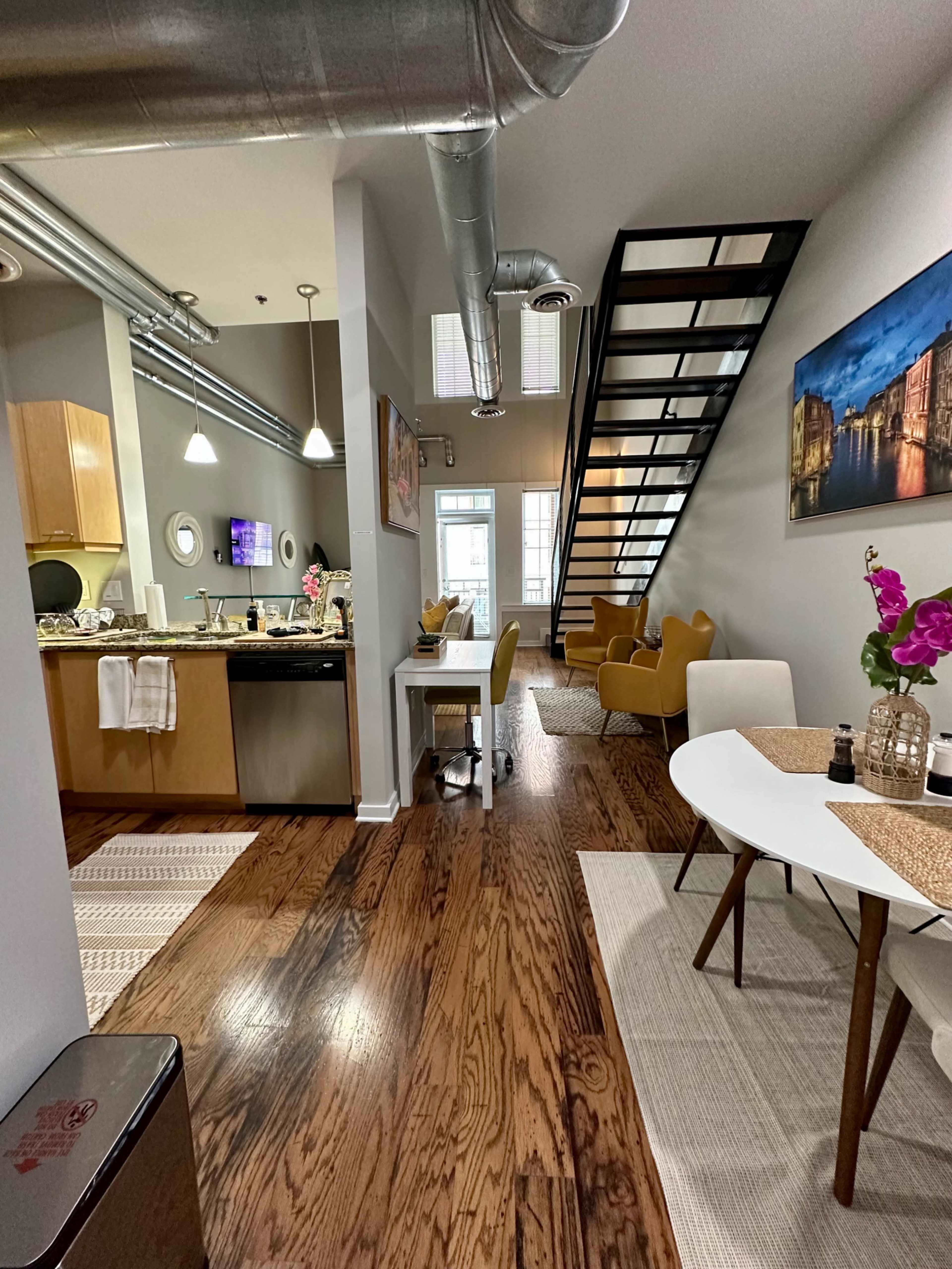 A spacious, modern apartment with a kitchen area to the left, a dining table and chairs to the right, and a staircase leading to a lofted area in the background.