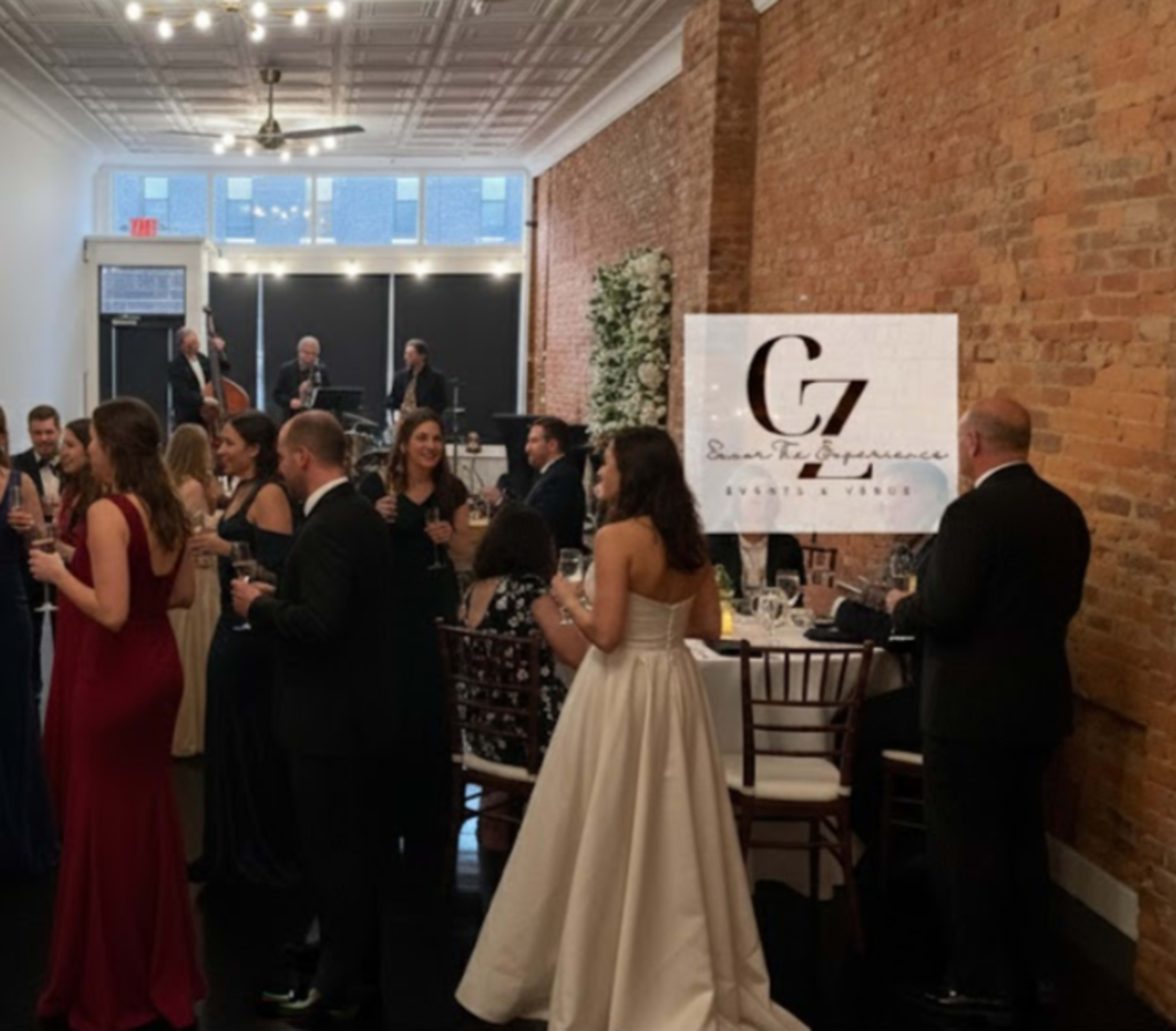 A group of guests in formal attire mingles at a stylish event in a venue with exposed brick walls and a live band performing in the background.