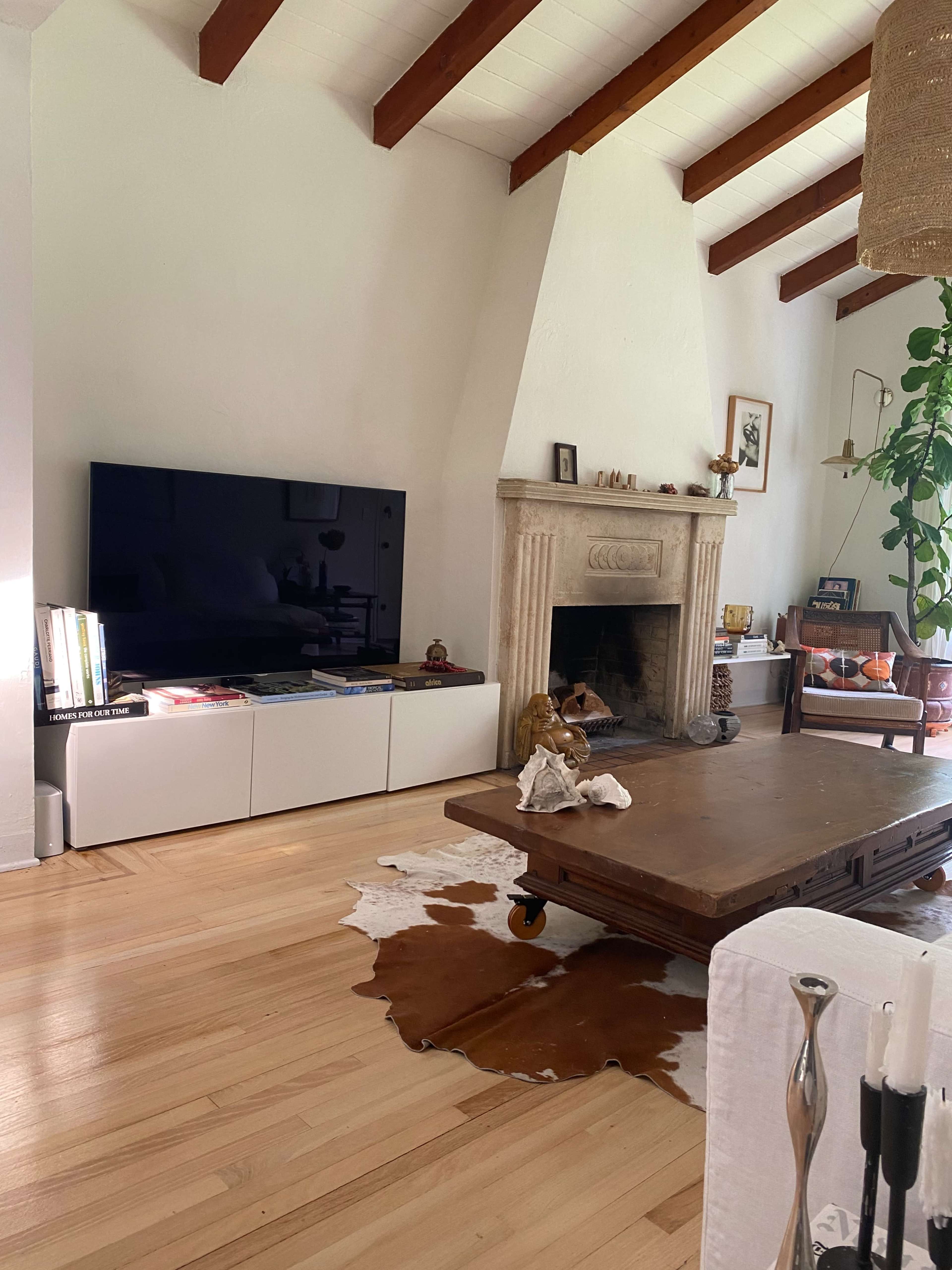 The image shows a living room with a wooden coffee table on a cowhide rug, a mounted television on a white cabinet, and a fireplace with decorative items.