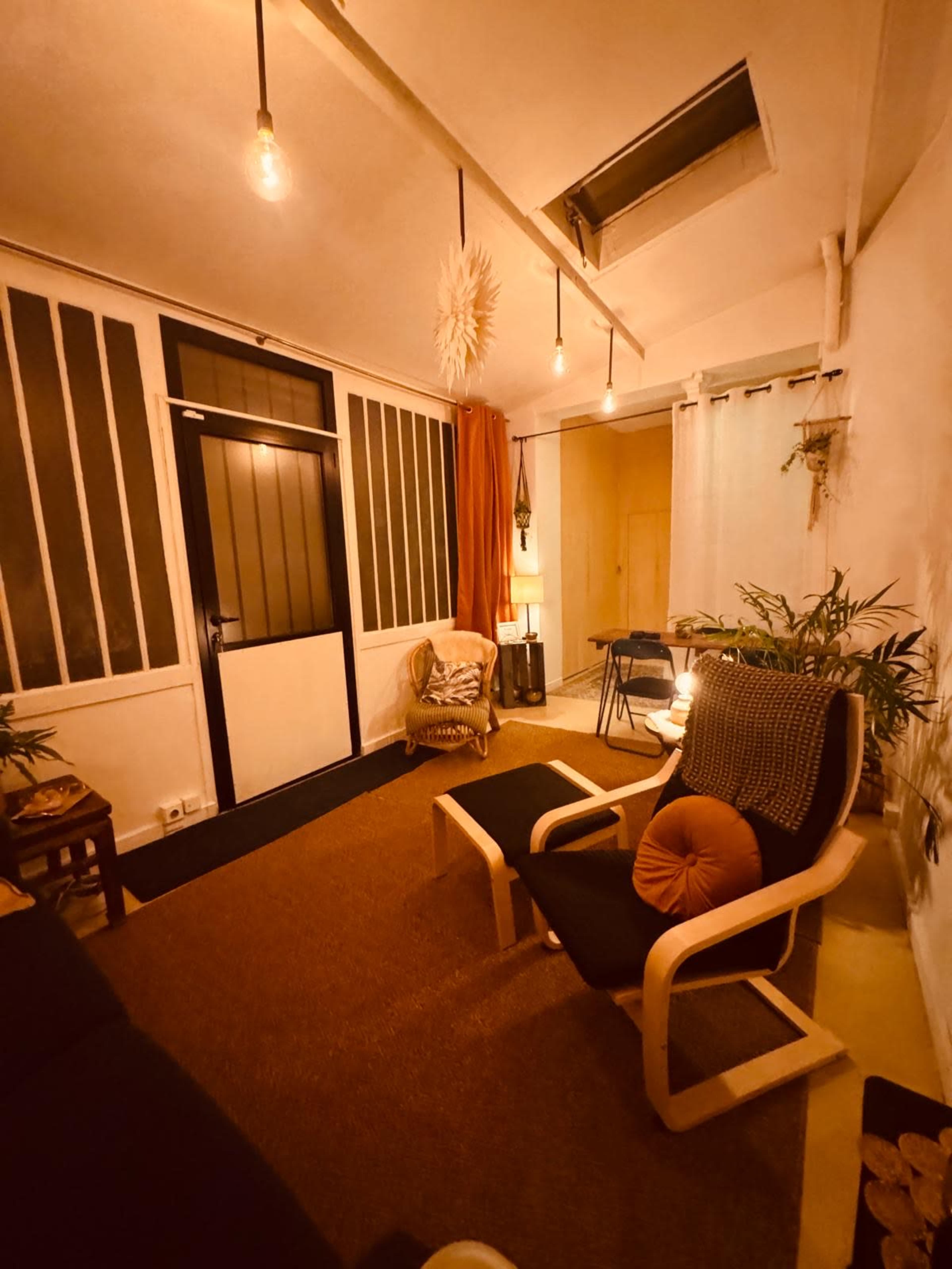 The image depicts a cozy interior space with a seating area, warm lighting, and various plants, featuring a door and large windows.