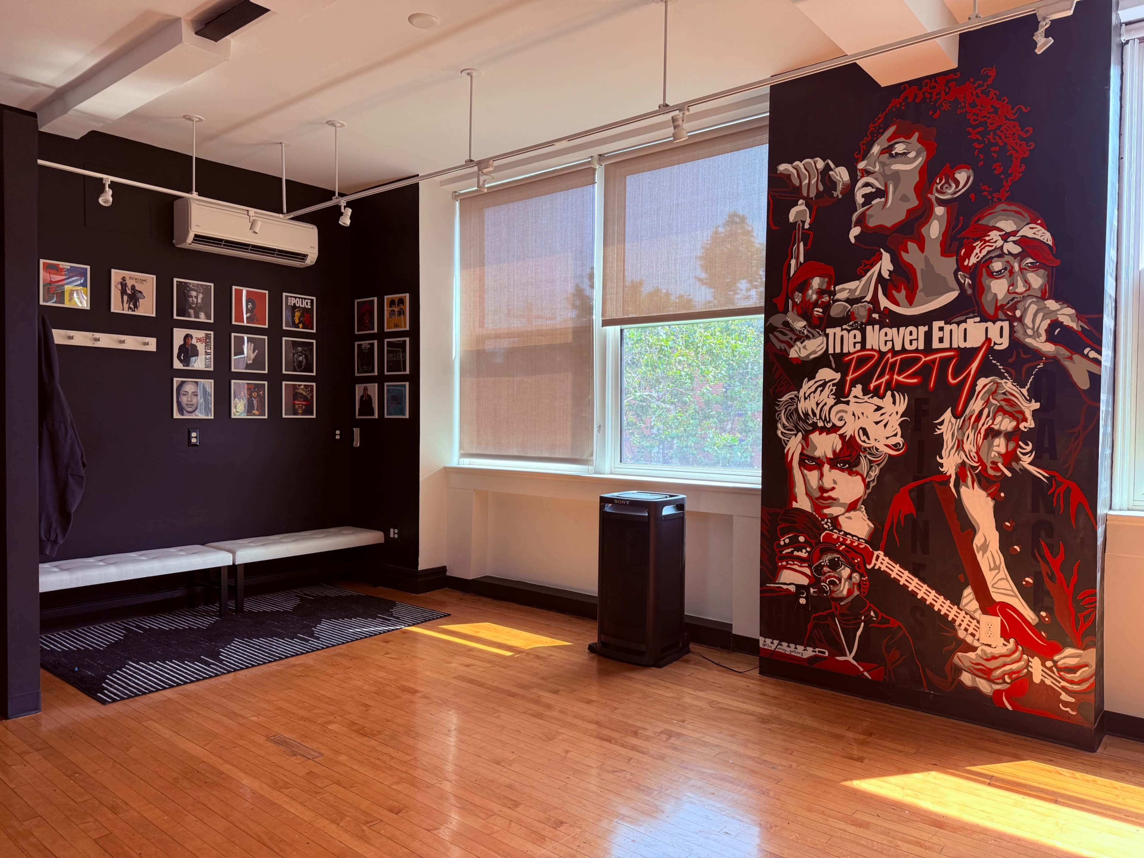 The image shows a room with dark walls, featuring a large mural of musicians, a bench along one wall, framed photographs, and a trash can near a window with sunlight streaming in.