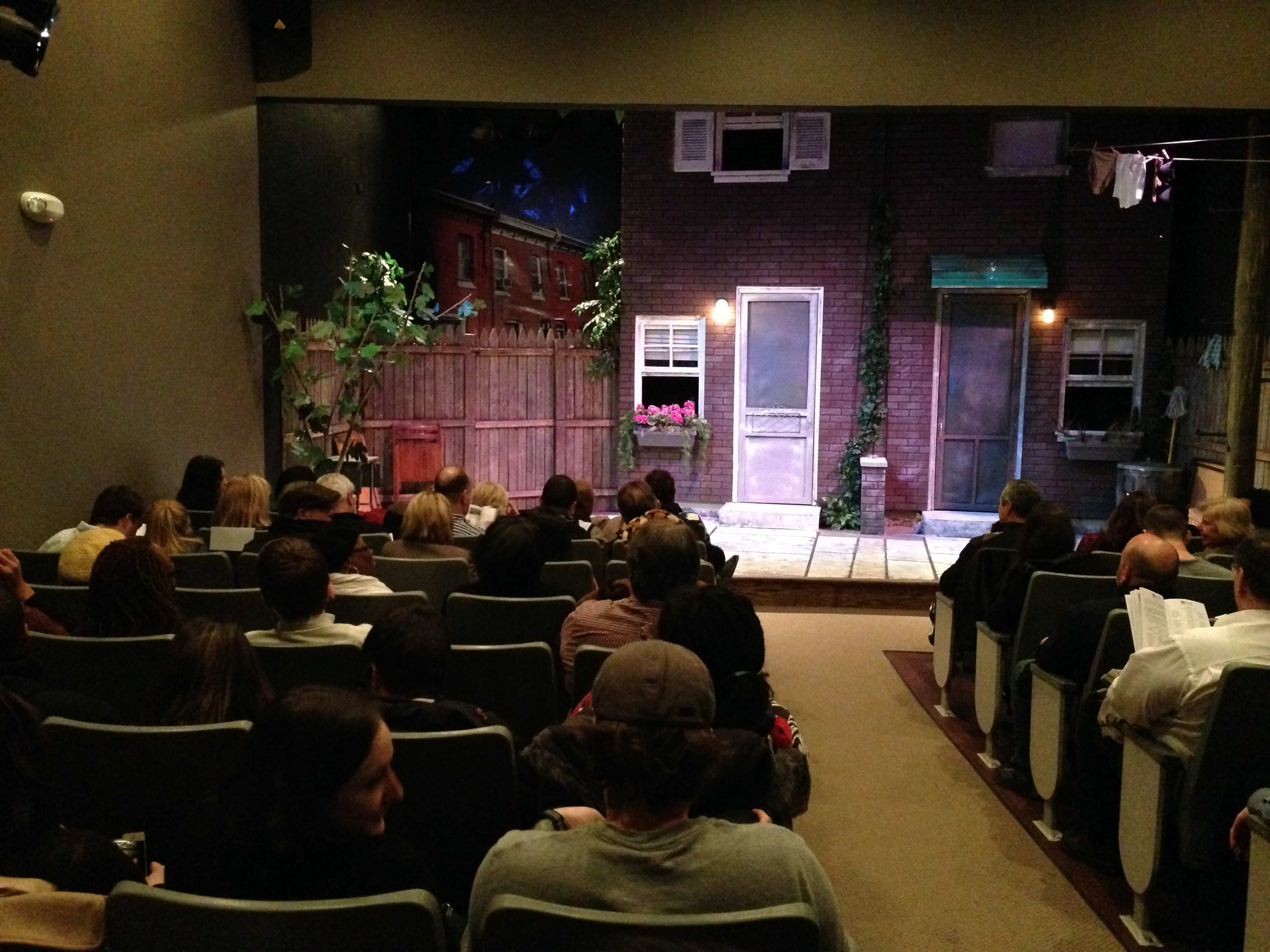 An audience sits in a small theater, facing a stage set designed to mimic a residential neighborhood with two doors and a fence.