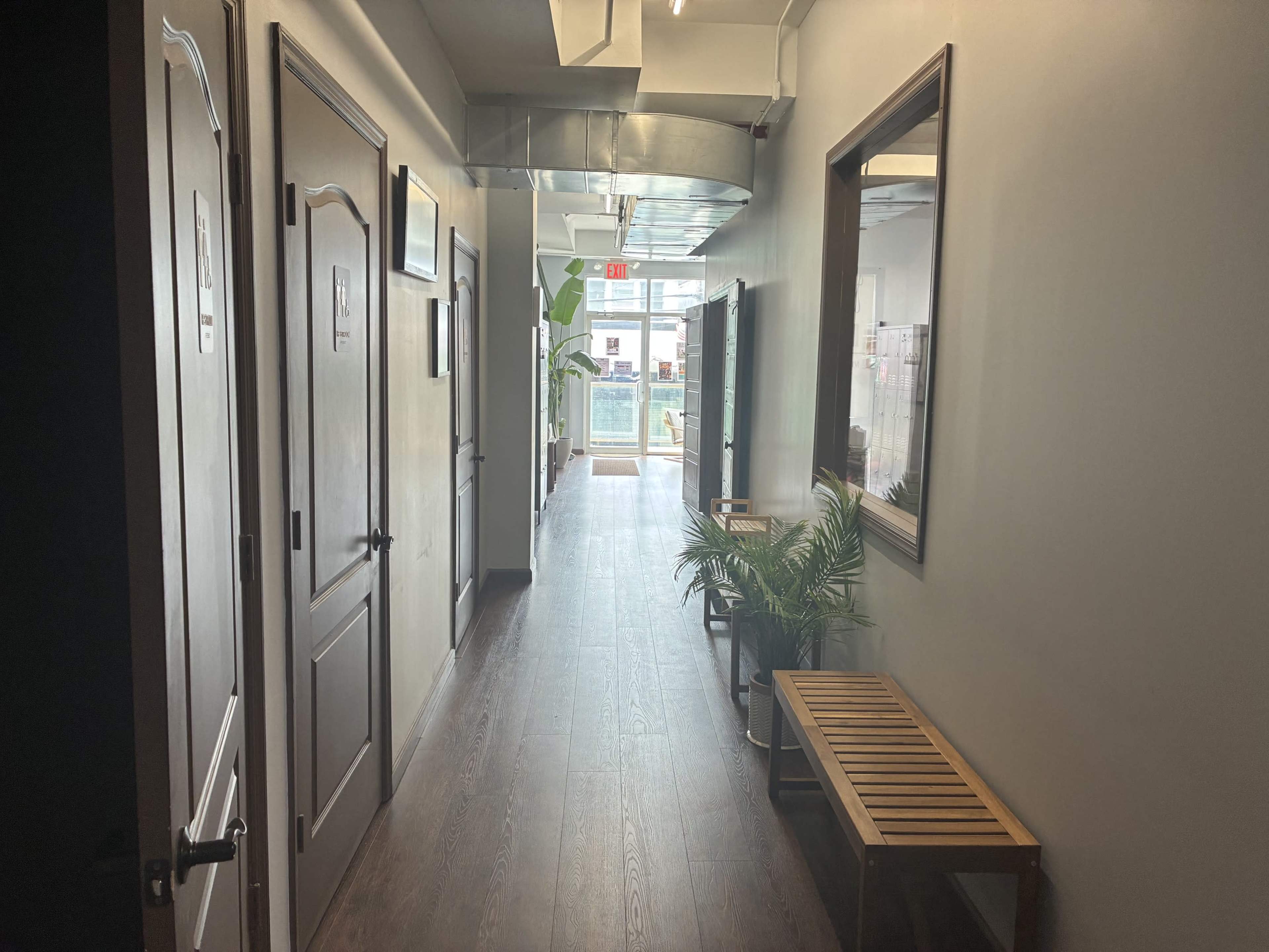 A hallway with closed doors on either side and a window at the end, featuring a bench and potted plants along the walls.