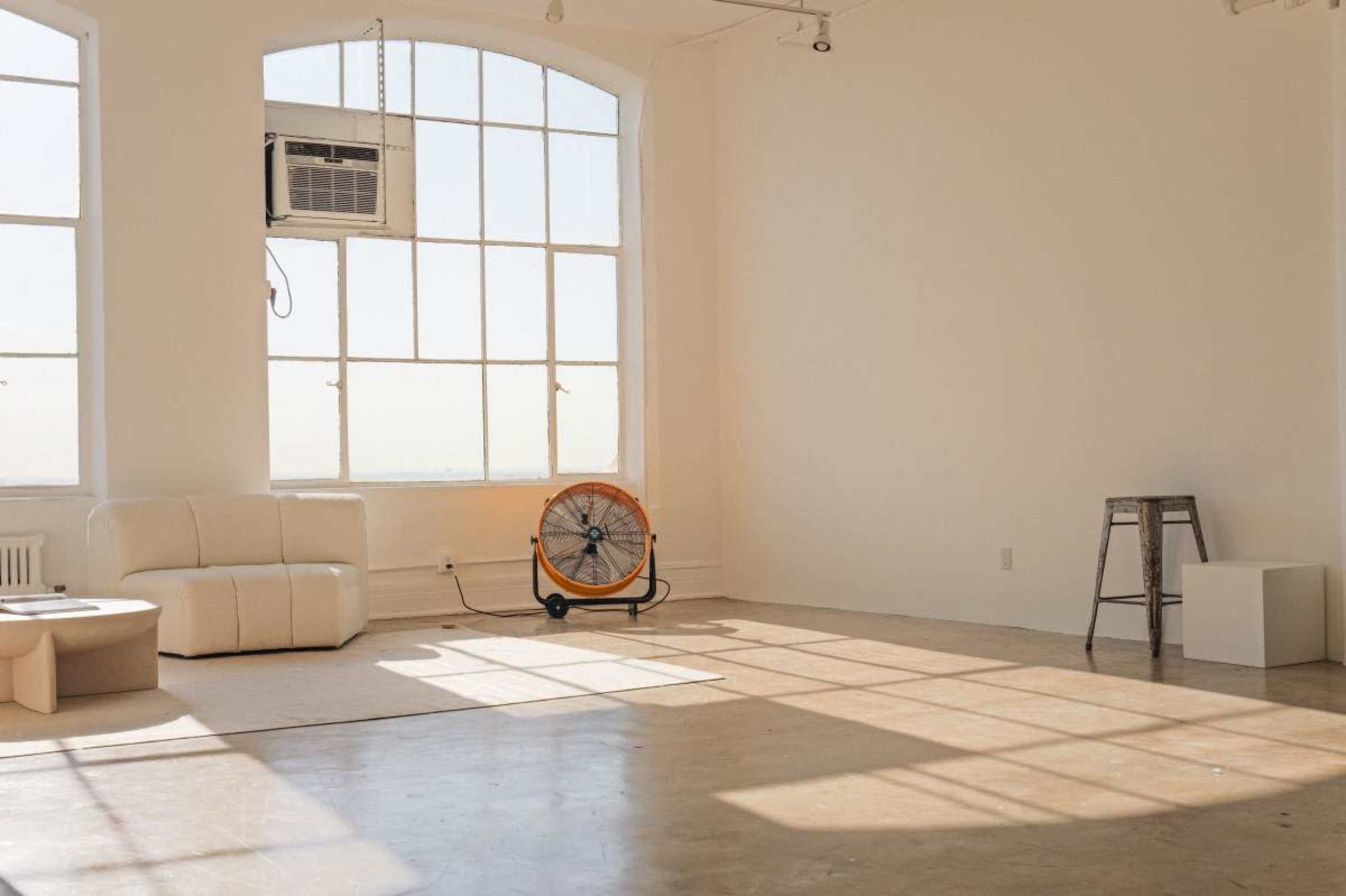 The image shows a spacious, well-lit room with large windows, a white couch, a fan, and a stool.