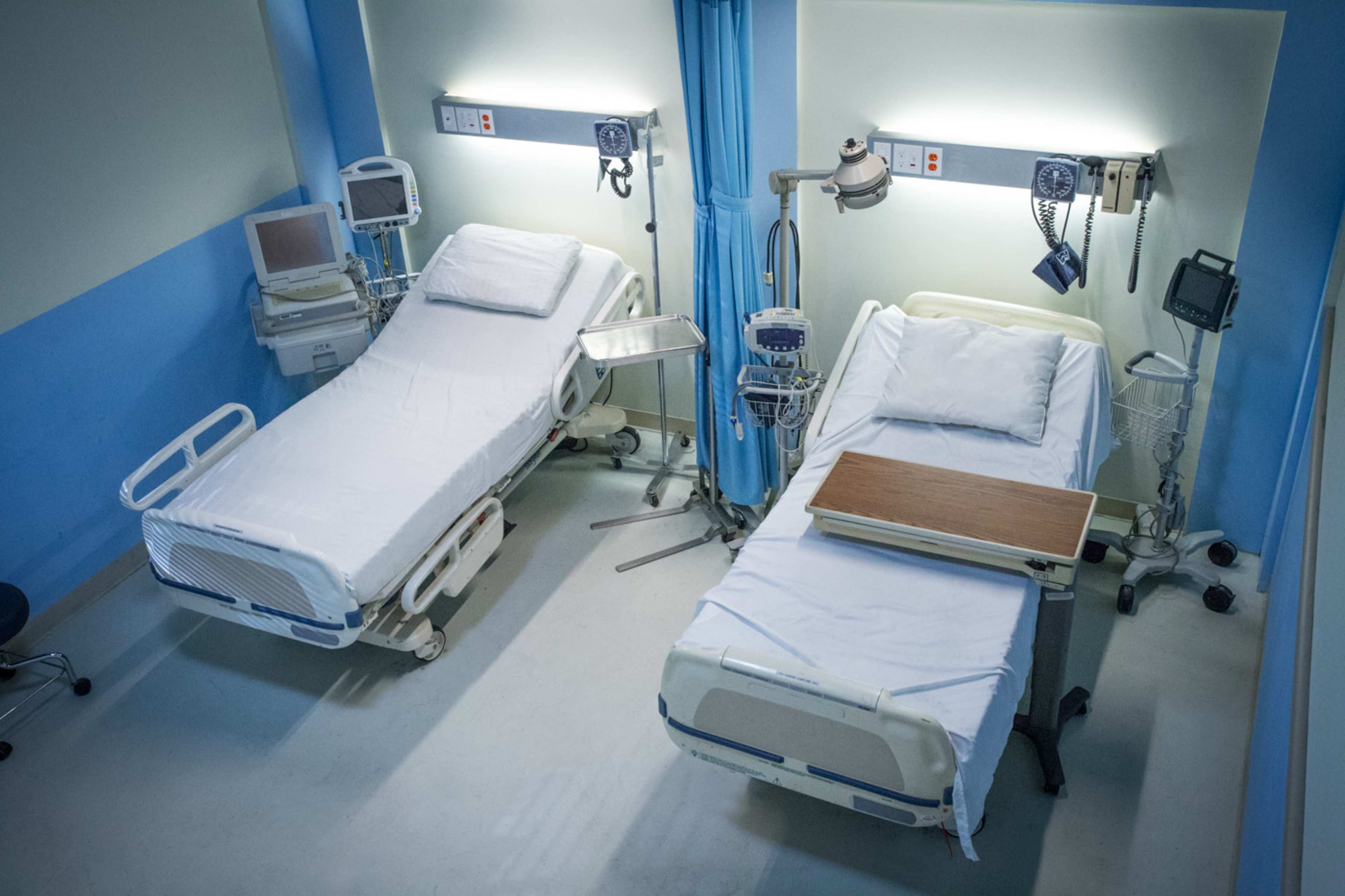 Two empty hospital beds in a brightly lit room, each with medical equipment nearby.