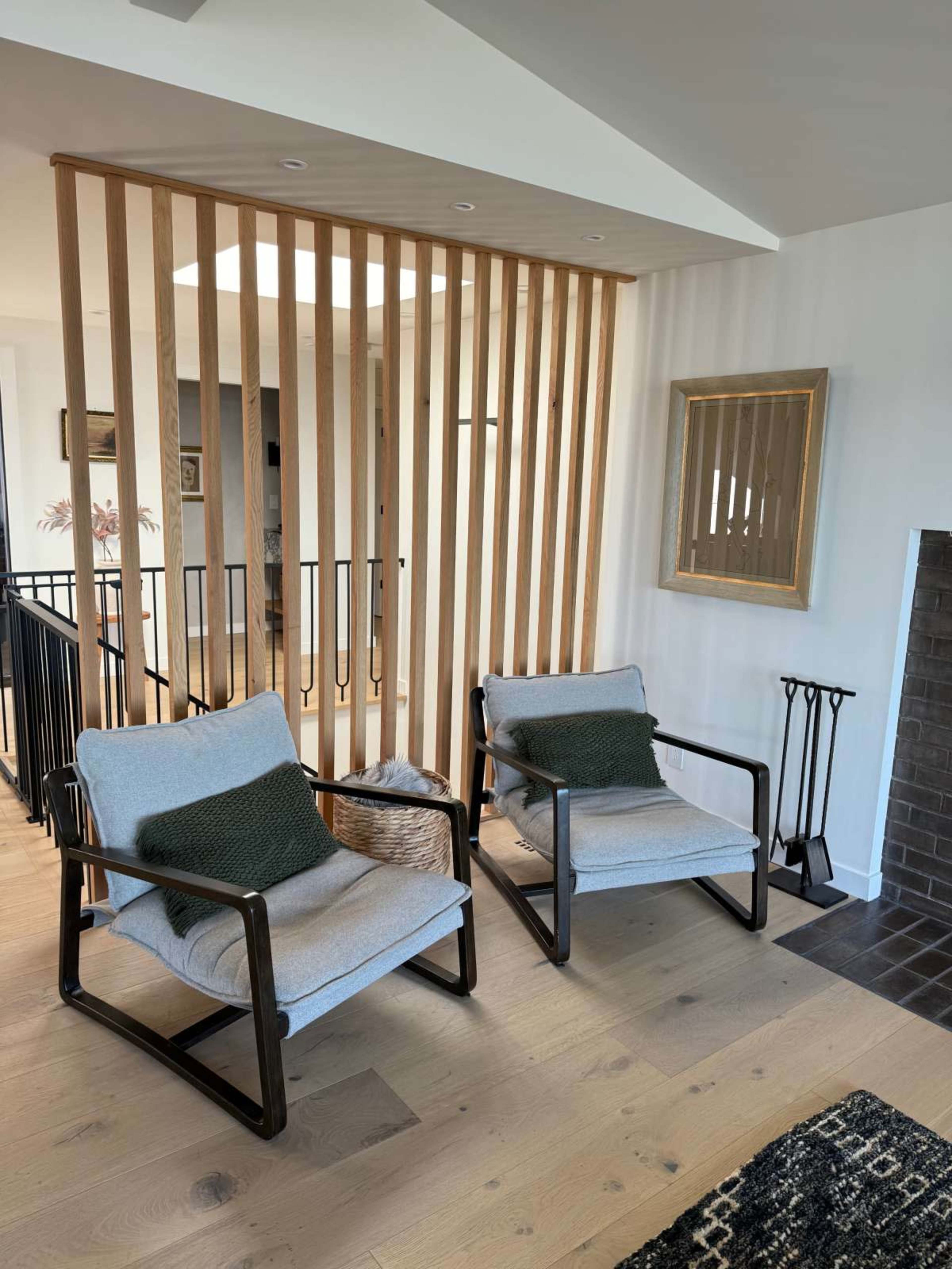 The image shows two modern chairs with cushions positioned on a wooden floor near a wooden slatted partition in a bright interior space.