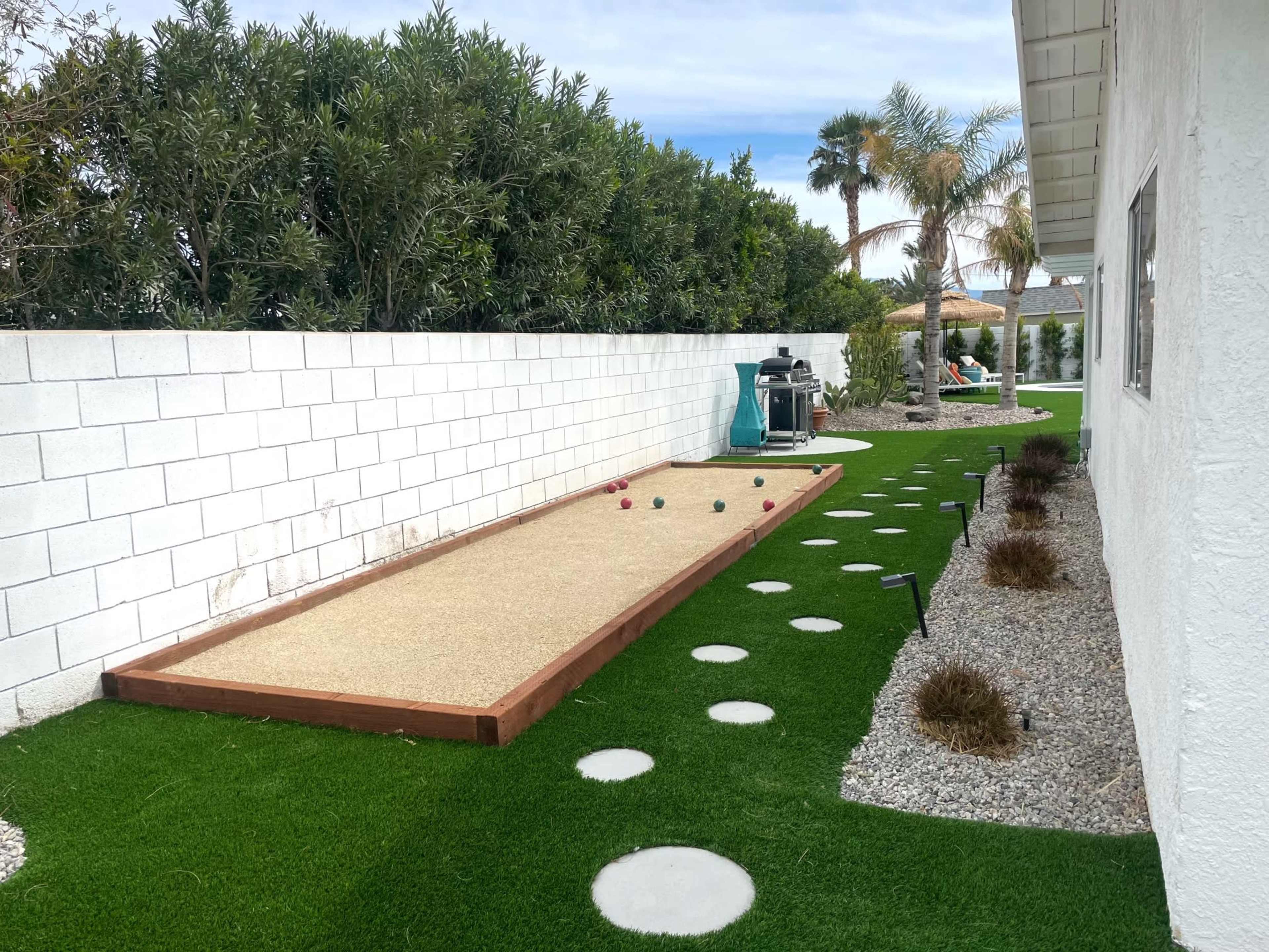 The image shows a bocce ball court bordered by a white wall, with artificial grass and decorative stones alongside it.