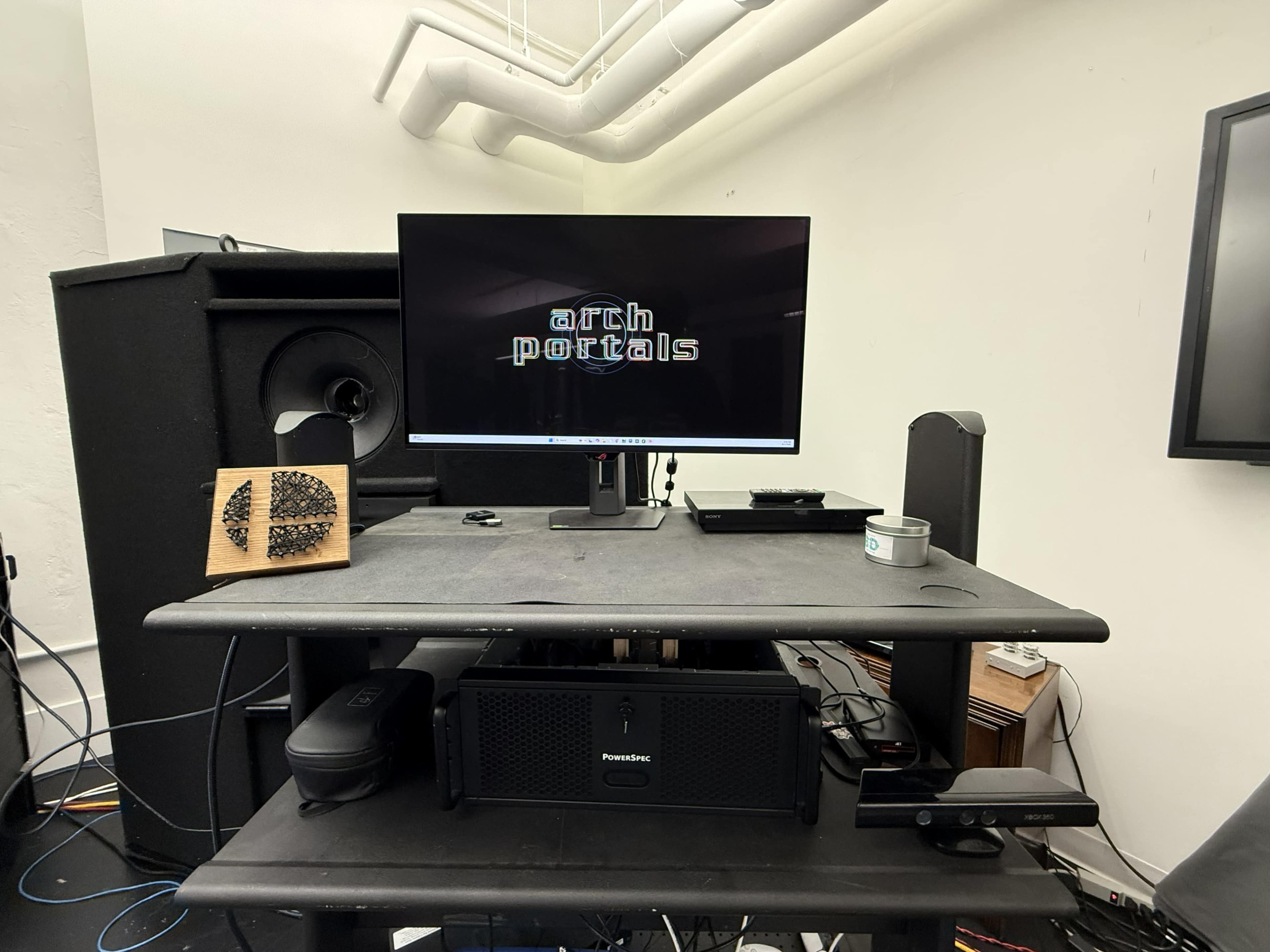A computer monitor displaying the text "arch portals" is positioned on a black desk, surrounded by various electronic devices and audio equipment.
