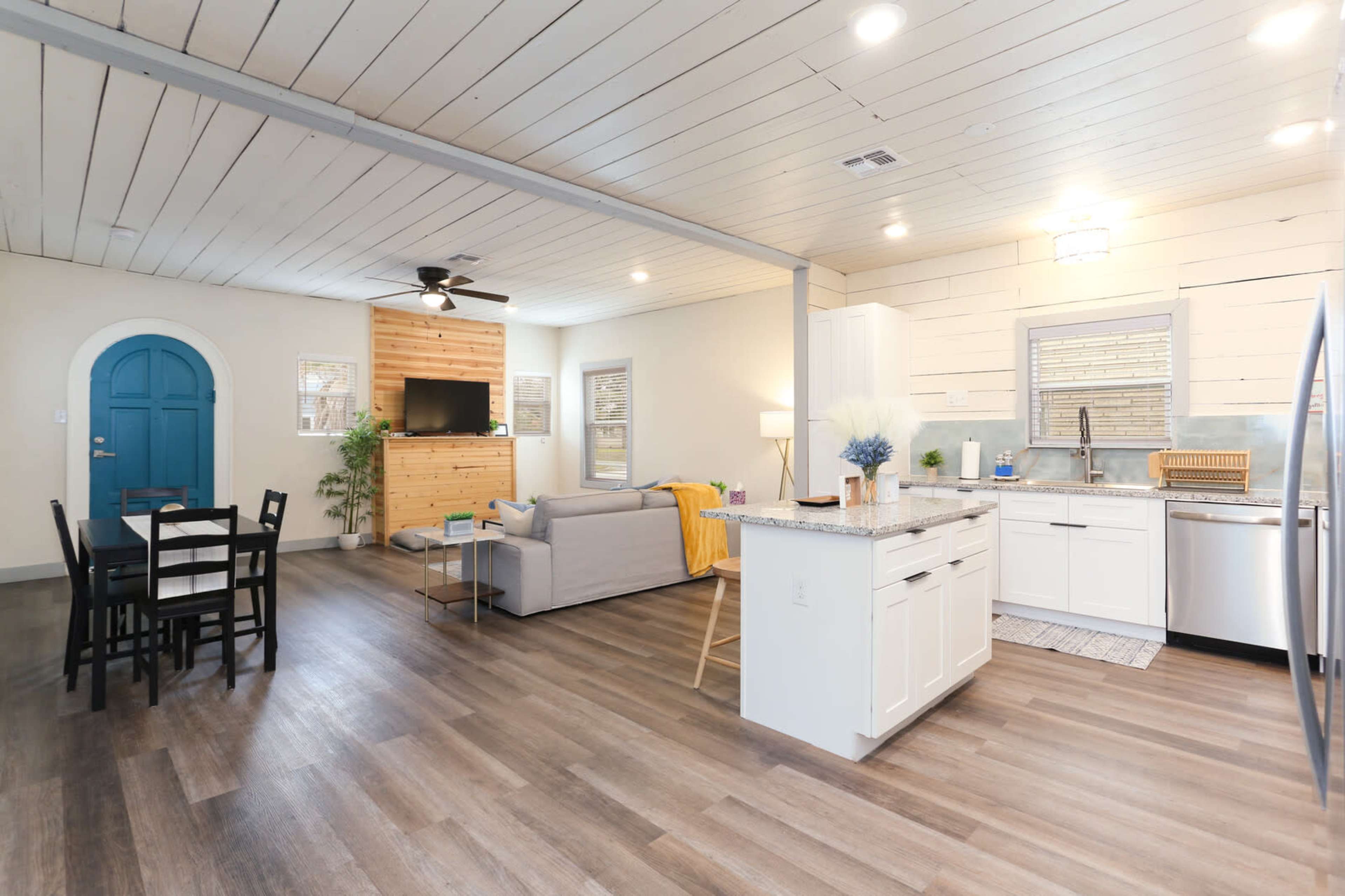 The image shows a modern open-concept living space with a kitchen, dining area, and a cozy seating area featuring a gray sofa and a television.