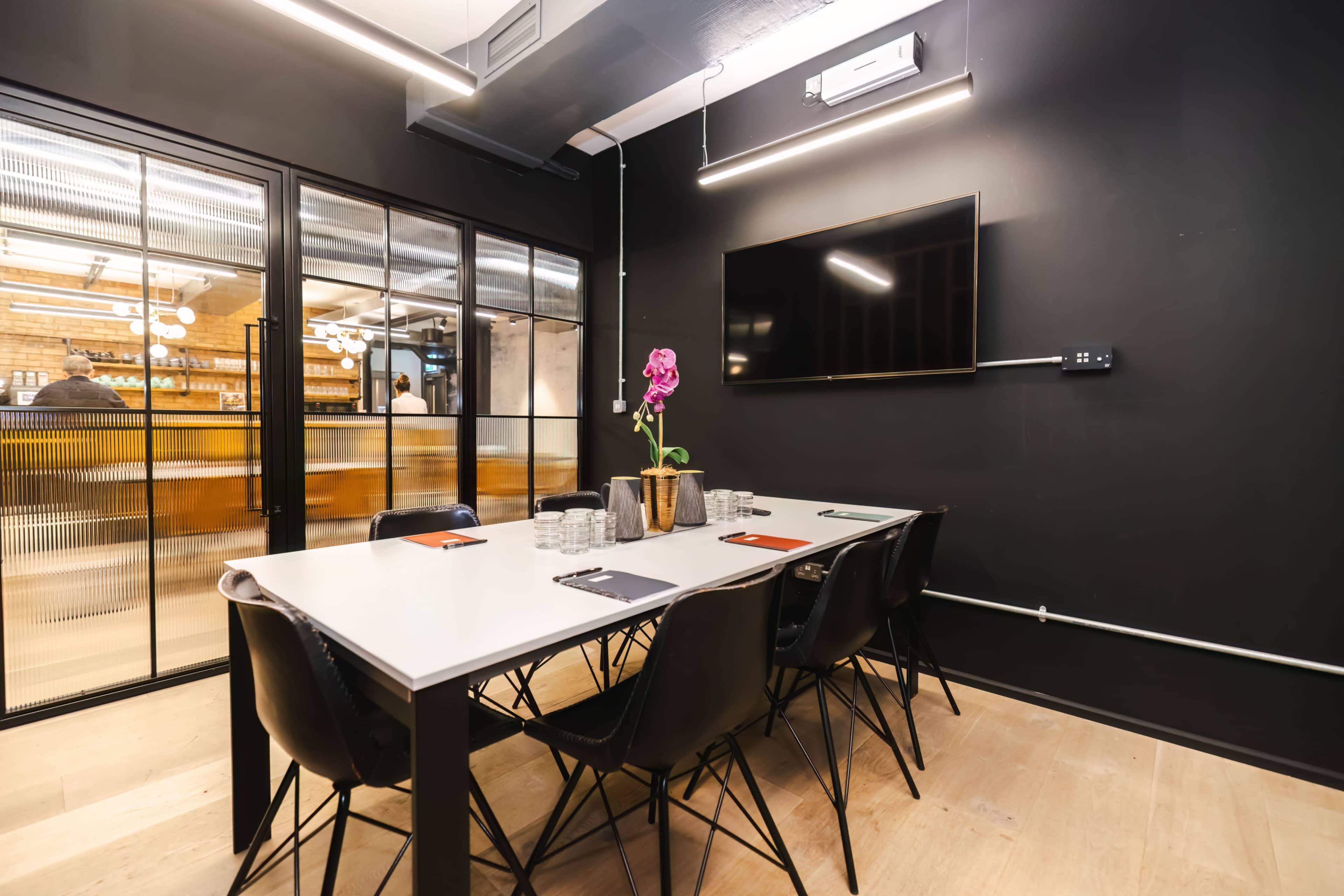 A meeting room featuring a white table surrounded by black chairs, with a television on the wall and a potted plant in the center, adjacent to a glass-walled area.