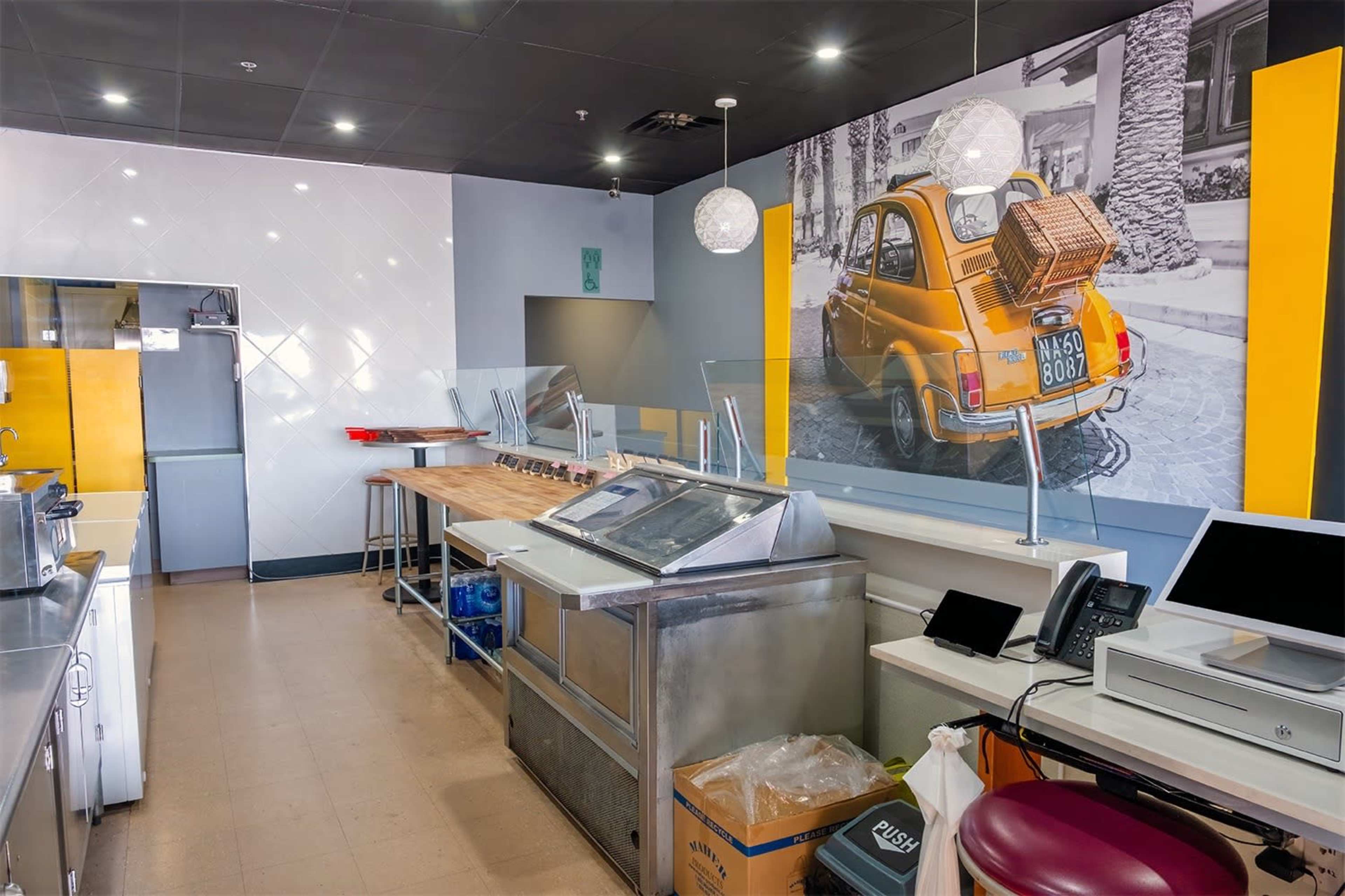 The interior of a small restaurant featuring a yellow vintage car mural on the wall, with kitchen equipment and a checkout station visible.
