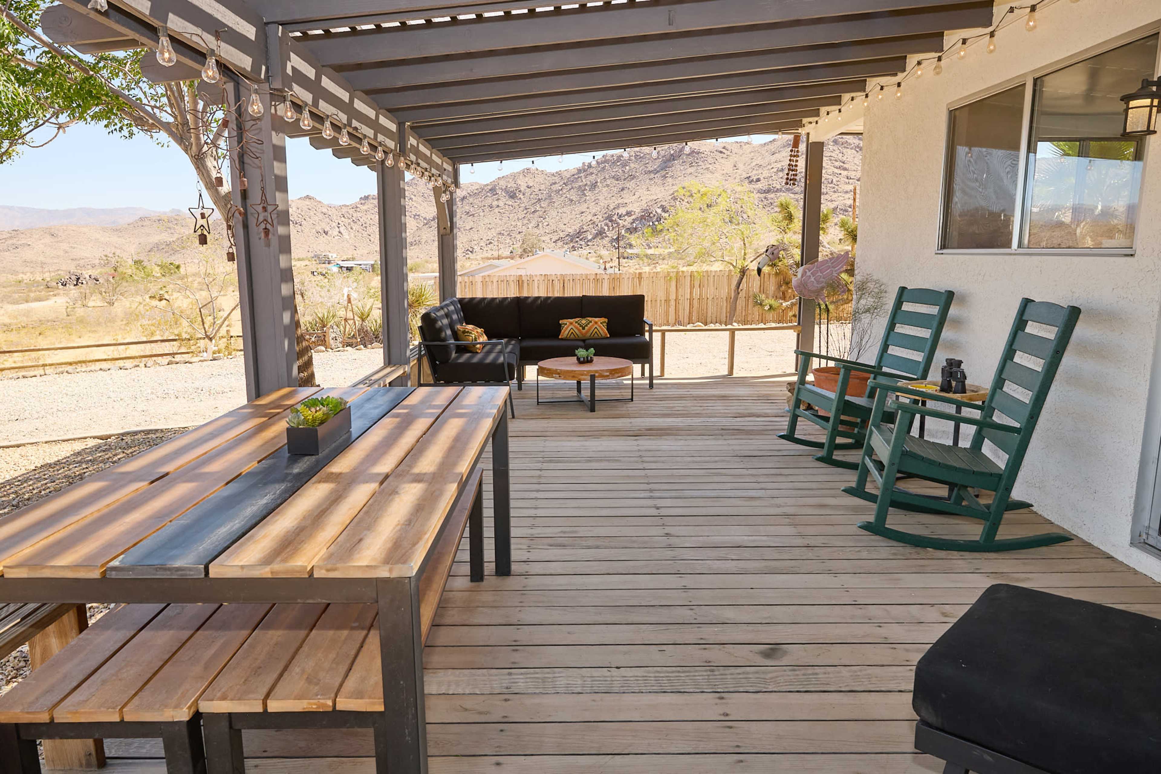 The image shows a patio area with a wooden dining table, rocking chairs, and a couch, surrounded by desert landscape and hills.