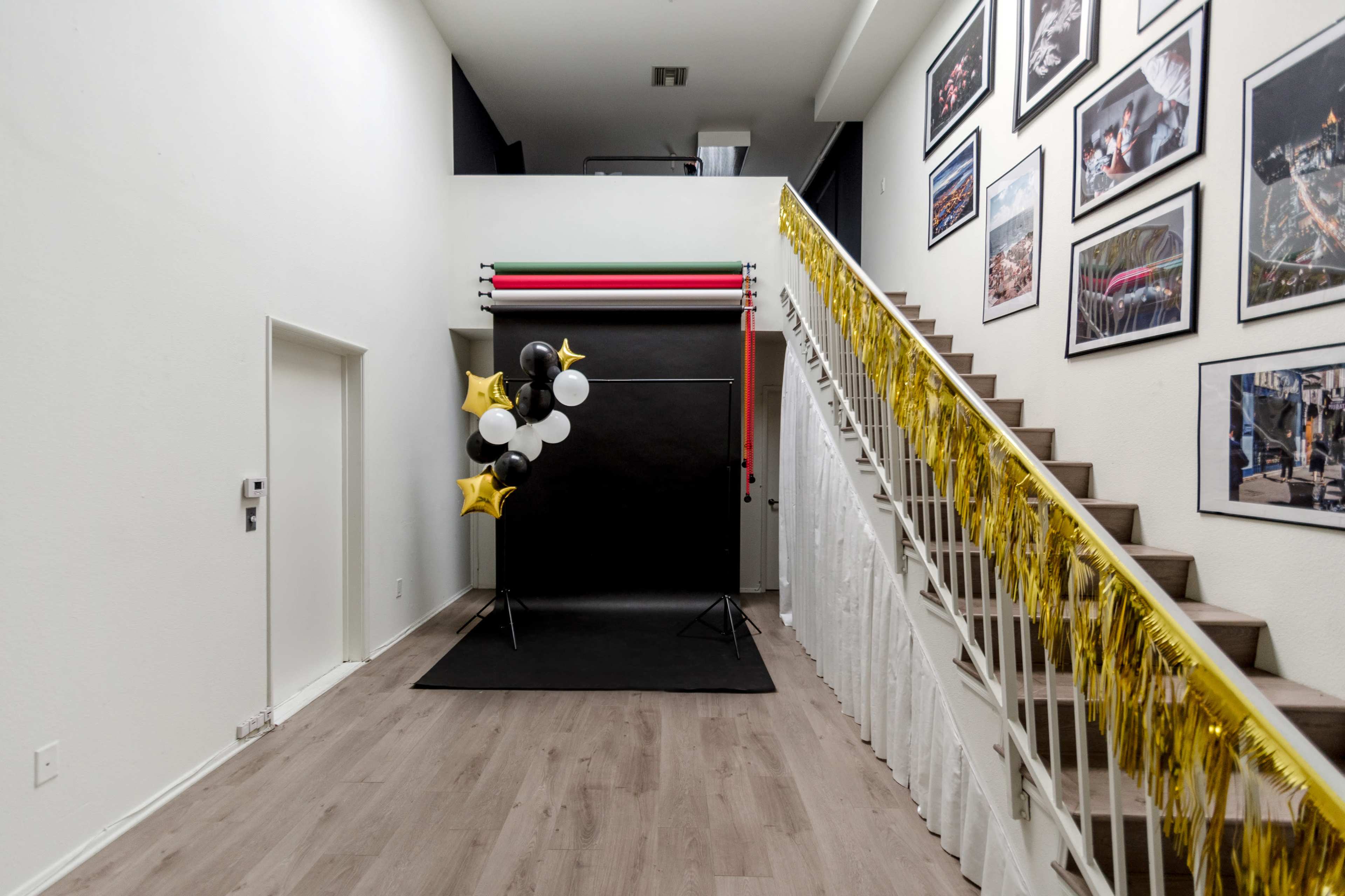 The image shows a well-lit hallway with a staircase, featuring a photo backdrop of black, white, and gold balloons, and walls adorned with framed photographs.